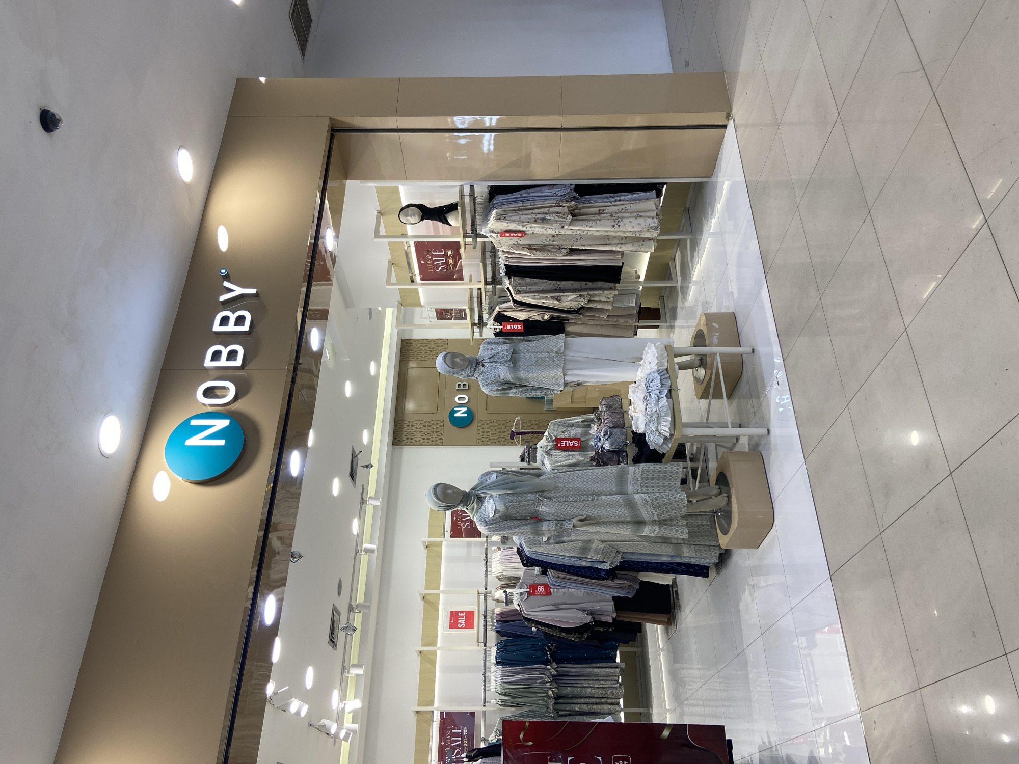 The image shows the exterior of a "NOBBY" clothing store in Kudus, Indonesia. The store has a beige facade with the "NOBBY" logo, featuring a blue circle with white lettering, prominently displayed above the entrance. The store entrance is a glass-fronted display showcasing mannequins dressed in modest fashion, predominantly long dresses and headscarves, in muted colors. Racks of clothing are visible inside, with several red "SALE" signs indicating promotions. The interior lighting is bright, illuminating the merchandise. The floor inside the store is tiled in white, while the surrounding area outside appears to be tiled in a light beige stone. The store is situated in what appears to be a shopping mall or commercial complex, given the indoor setting and the presence of ceiling lights and what looks like a security camera.