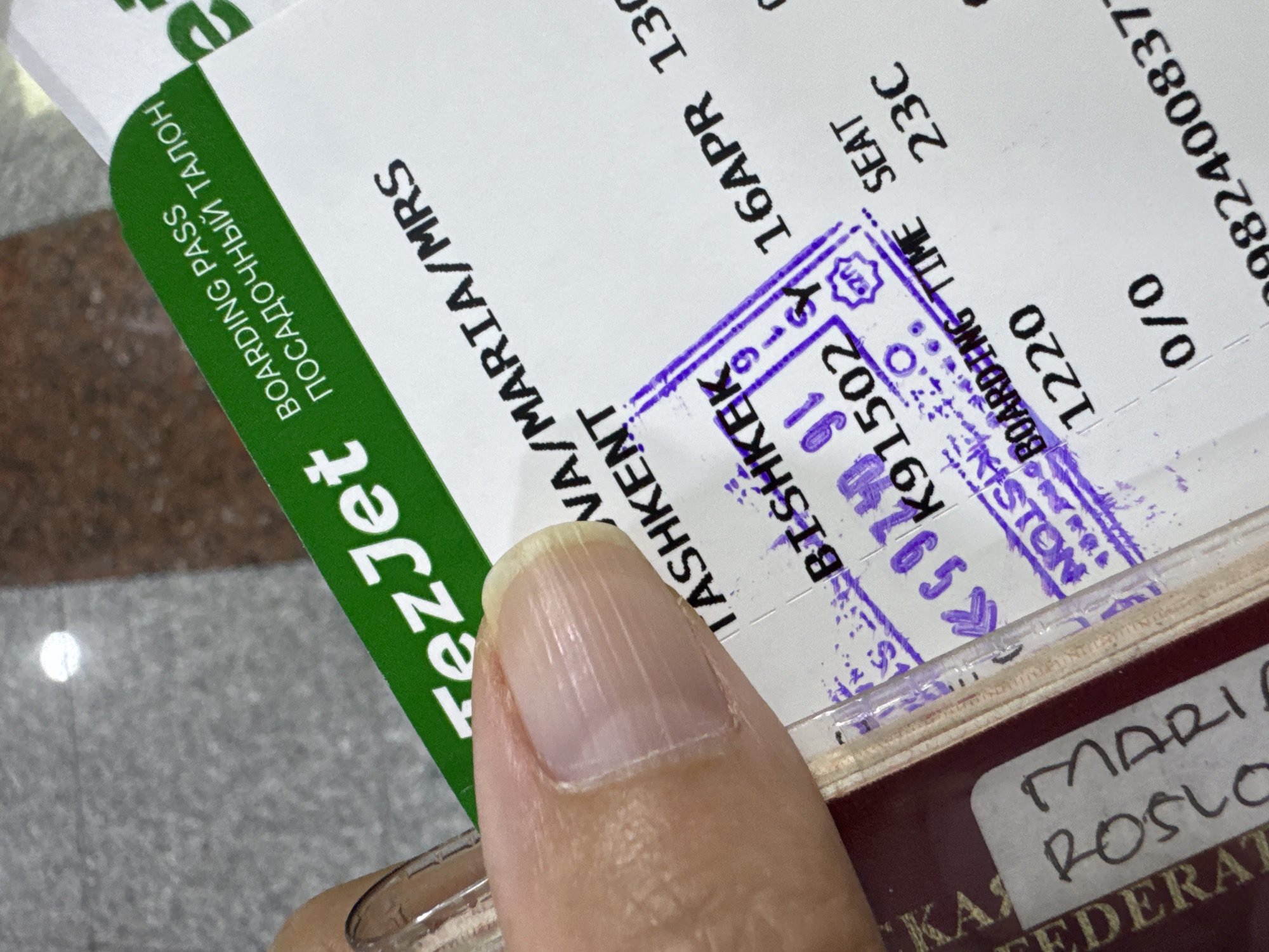 A boarding pass issued by TezJet with "BOARDING PASS ПОСАДОЧНЫЙ ТАЛОН" printed in green on the left side is shown. The boarding pass is for a flight from Tashkent (TAS) to Mineralnye Vody (MRS) for a passenger with the name "VA/MARIA/MRS" on April 16. The seat number is 23C, and the boarding time is 1220. A purple ink stamp with a date and code "K91502" is visible on the boarding pass over "BISHKEK," suggesting it was processed in the city of Bishkek. A passport is visible under the boarding pass. The passport contains the name "MARIA ROSLO" and the words "FEDERAT KAS". The scene is indoors. The image is partially obscured by a person's thumb.
