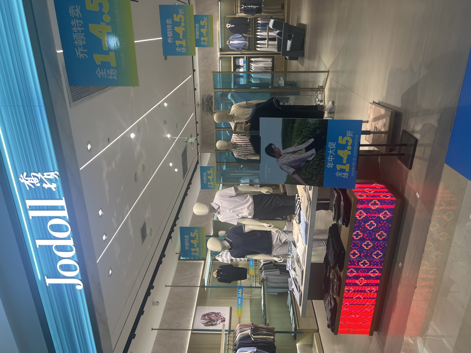 The image shows the interior of a Jodoll clothing store, likely located in Liandu District, China, as indicated by the store name and prominent signage. The store features a modern design with bright lighting and display shelves filled with men's clothing. Two mannequins are prominently displayed in the foreground, one wearing a striped polo shirt and the other a light-colored collared shirt. They are positioned in front of a table displaying folded shirts. In the background, more clothing is visible on racks and shelves, including jackets, shirts, and pants. Several promotional signs are hung from the ceiling, advertising a sale with discounts ranging from 1 to 4.5, with dates suggesting the sale period is from July 1 to August 31, 2026. A poster featuring a young man in a casual outfit is also visible near the mannequins. The floor is tiled and reflective, with shadows cast by the lighting and displays.