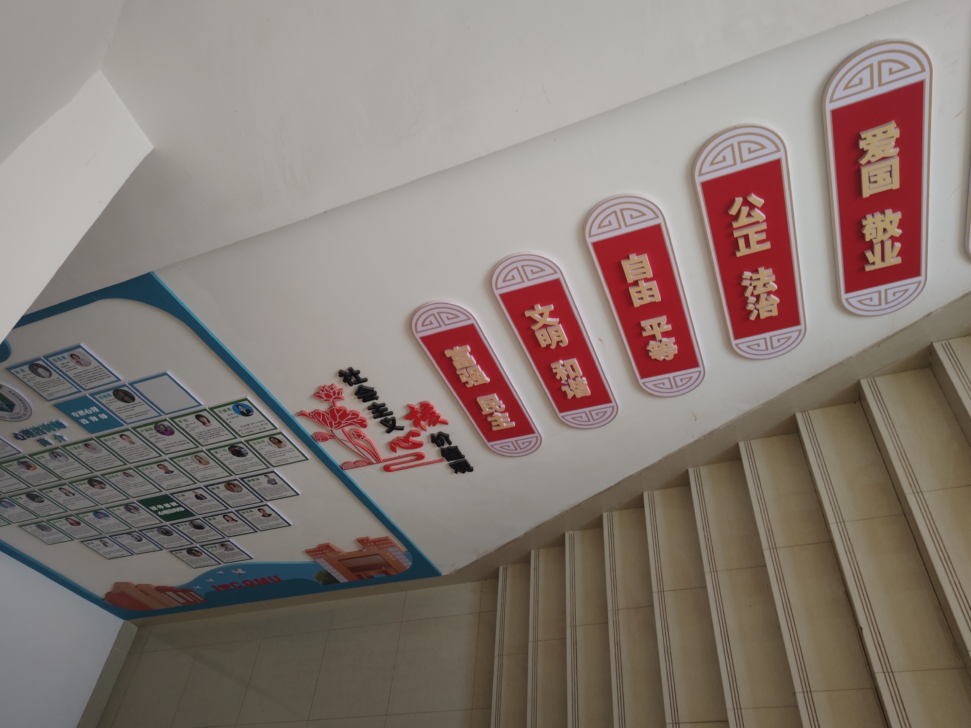 The image shows a staircase in what appears to be a public building or school located in the city of Chongqing High-tech Zone, China. The stairs are lined with cream-colored tiles and feature thin, dark grooves along the edges of each step for better grip. On the wall above the staircase, various decorative elements are displayed. These include red, stylized plaques with Chinese characters, arranged in descending order of size from right to left. From right to left, the text on the plaques, transliterated to English, reads: 'Love Country Respect Occupation', 'Justice Rule', 'Freedom Equality', 'Civilized Harmony' and 'Rich Strong Democracy'. To the left of the plaques, a bulletin board shows numerous small photo identification cards, seemingly displaying headshots and names of individuals. The Chinese text '心理咨询师' (Psychological Counselor) appears on one of the cards. Overall, the decorations suggest a focus on civic values, education, and psychology.
