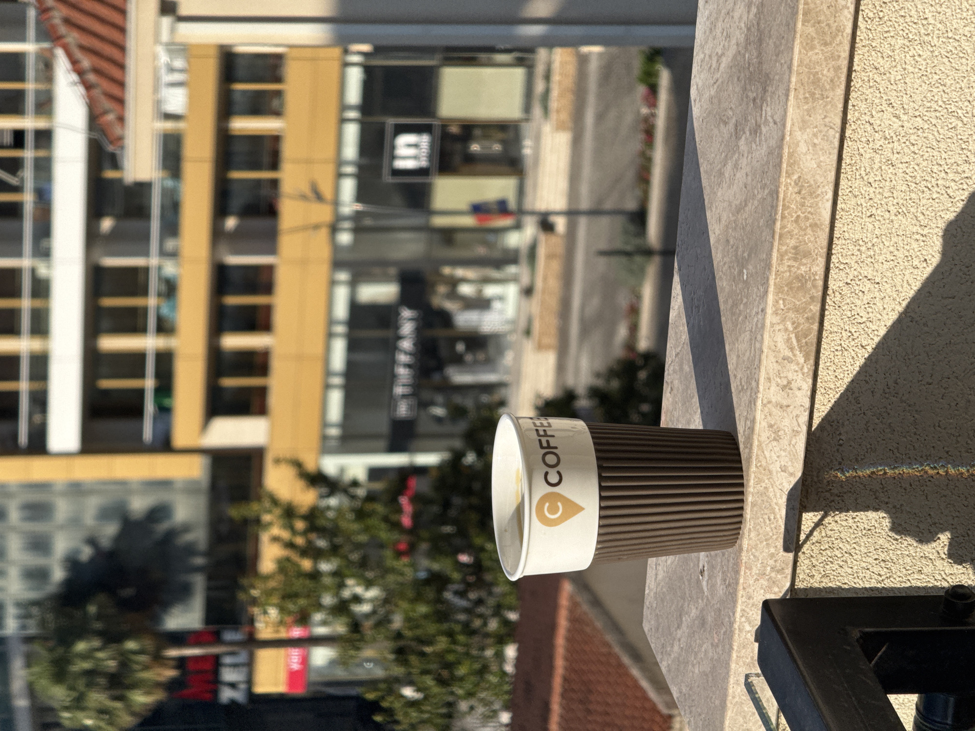 A close-up, eye-level shot captures a brown ribbed paper coffee cup filled with a light-colored liquid, possibly coffee, resting on a marble ledge. The cup features a white sleeve with a brown logo containing the text "C COFFEE" and a small icon of a map pin. The background is blurred, showing a modern building with yellow accents and balconies, indicative of a city environment. Signs for "TIFFANY" and "IN STORE" are visible on the building's storefront. A palm tree and other greenery add to the urban landscape. Sunlight casts a shadow from the cup onto the textured wall below, with a subtle rainbow effect visible on the wall. The scene is likely from Budva, Montenegro, given the architectural style and prominent palm tree.