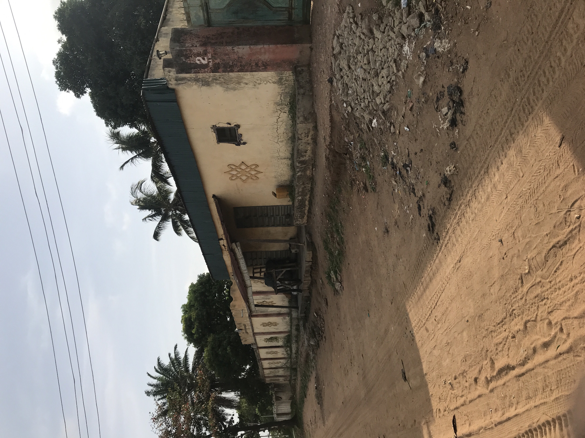 The image shows a street scene in Oko Erin, Nigeria. A weathered, single-story building with a pale yellow facade stands adjacent to a dirt road. The building has a green metal roof, a small square window, and decorative geometric patterns on the wall. The address "2 YOLA RD." is visible on the building's front. A small covered porch area is supported by posts.
The road is sandy with visible tire tracks. Debris and loose rocks are scattered along the roadside. Trees, including palm trees, are visible along the street, with power lines running overhead. The sky is overcast. There are no visible people or signs of recent human activity.