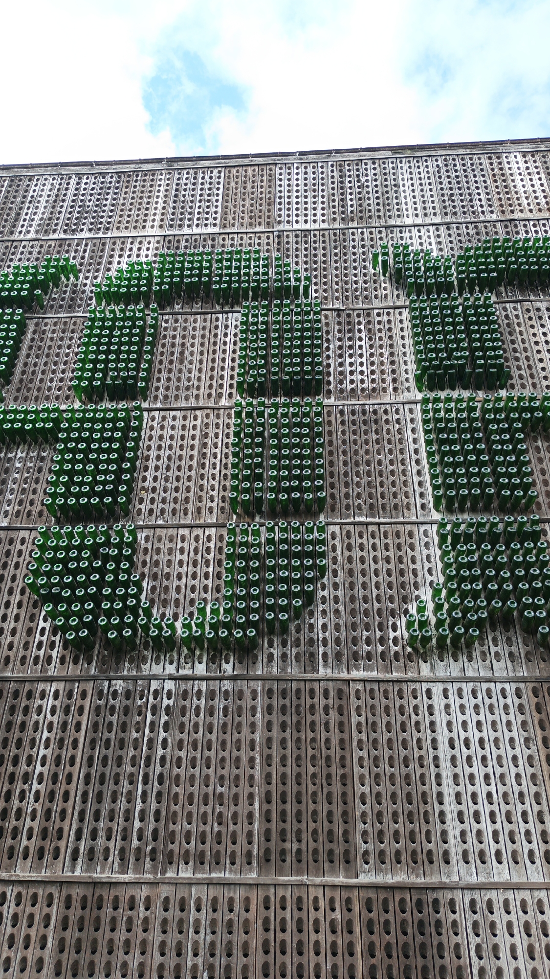 Here's a description of the image:
The image shows a wall facade in the city of Abrau-Dyurso, Russia. The wall is constructed from vertical, wooden panels with rows of circular holes. Green glass bottles have been inserted into some of the holes to form a decorative pattern, spelling out the name "ABRAU". The sky is visible in the top part of the image.