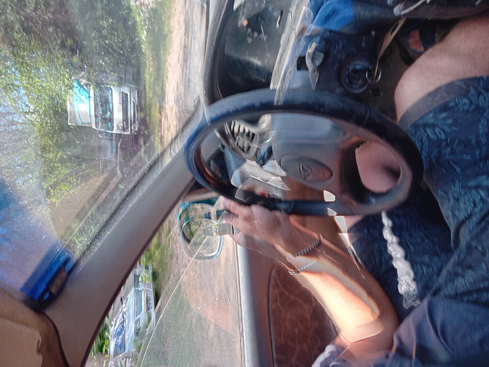 The image shows the interior of a car, with a person's hands on the steering wheel. The car appears to be moving, as evidenced by the motion blur. The driver is wearing a short-sleeved shirt and shorts. Outside the car, there are trees and a truck visible. Based on the context provided by the file name, this scene is likely from Chuhuiv, Ukraine.