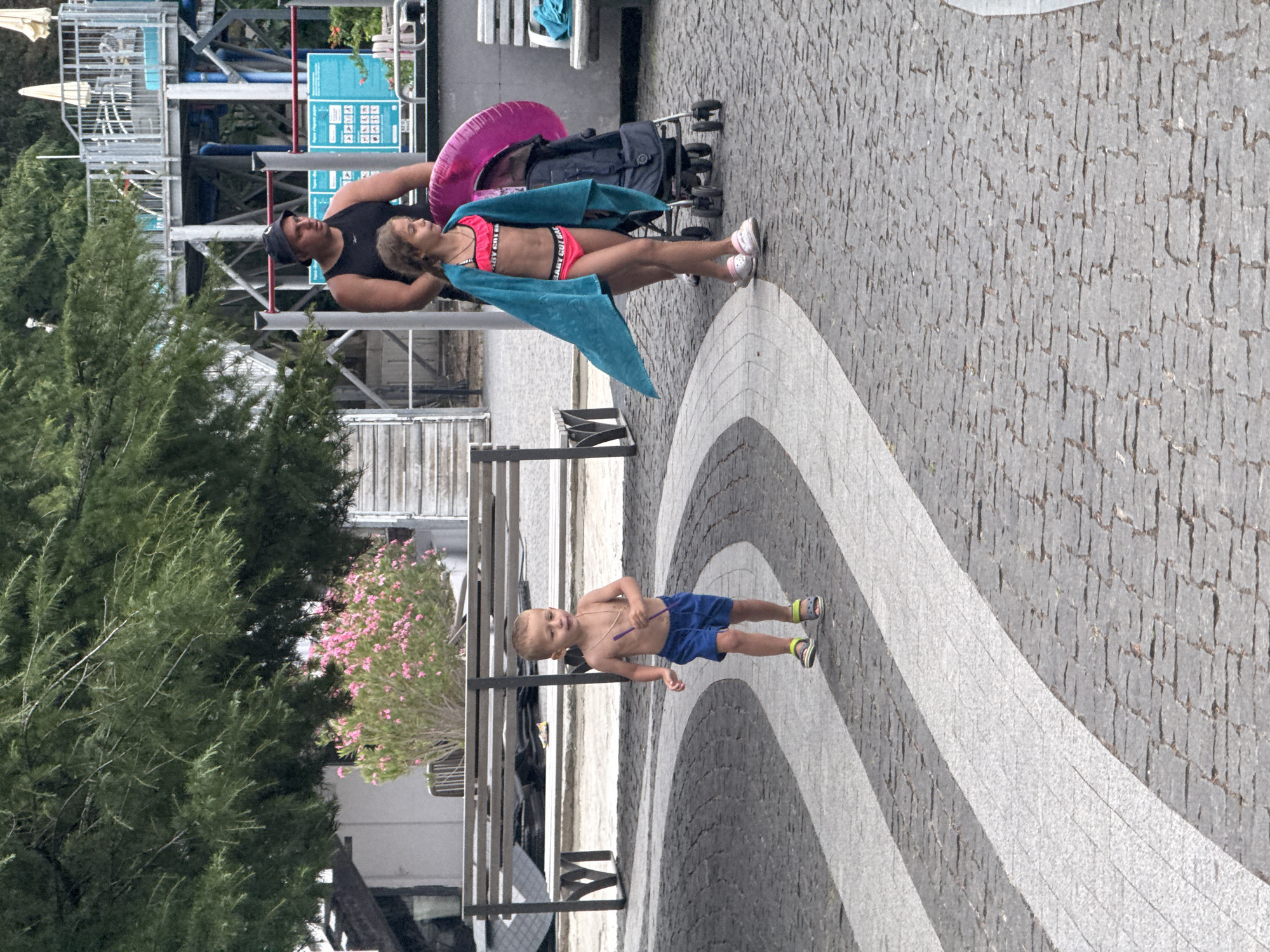 The image depicts a family enjoying a day at an outdoor water park or similar recreational facility in Yalta, Ukraine. In the foreground, a young boy with blond hair, wearing blue swim trunks and sandals, stands on a cobblestone path. He is holding a purple stick and looking towards the camera. Behind him, a teenage girl in a pink bikini and a blue towel draped over her shoulders stands with a man in a black tank top. They are next to a stroller with a pink inflatable ring. The background shows water park attractions, including slides and metal structures, along with trees and bushes, some of which are in bloom with pink flowers. The scene appears to be set during daylight, with overcast skies suggesting potentially cooler weather or preparation for rain. The overall atmosphere seems relaxed and familial, with the subjects appearing engaged with each other or the surroundings. Visible text includes "BABY GOT BACK" on the girl's bikini bottoms.