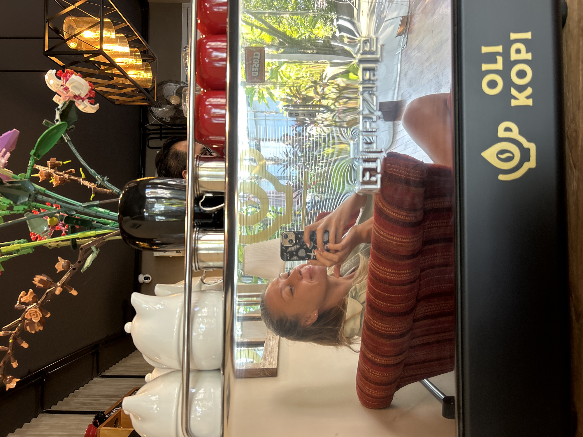 The image shows a reflection of a woman taking a selfie in the polished surface of a coffee machine. She is sitting in a red striped armchair. The coffee machine has a logo that says "OLI KOPI". In the background, there are plants visible through a window and a "CLOSED" sign. The scene appears to be a cafe in North Kuta, Indonesia.
