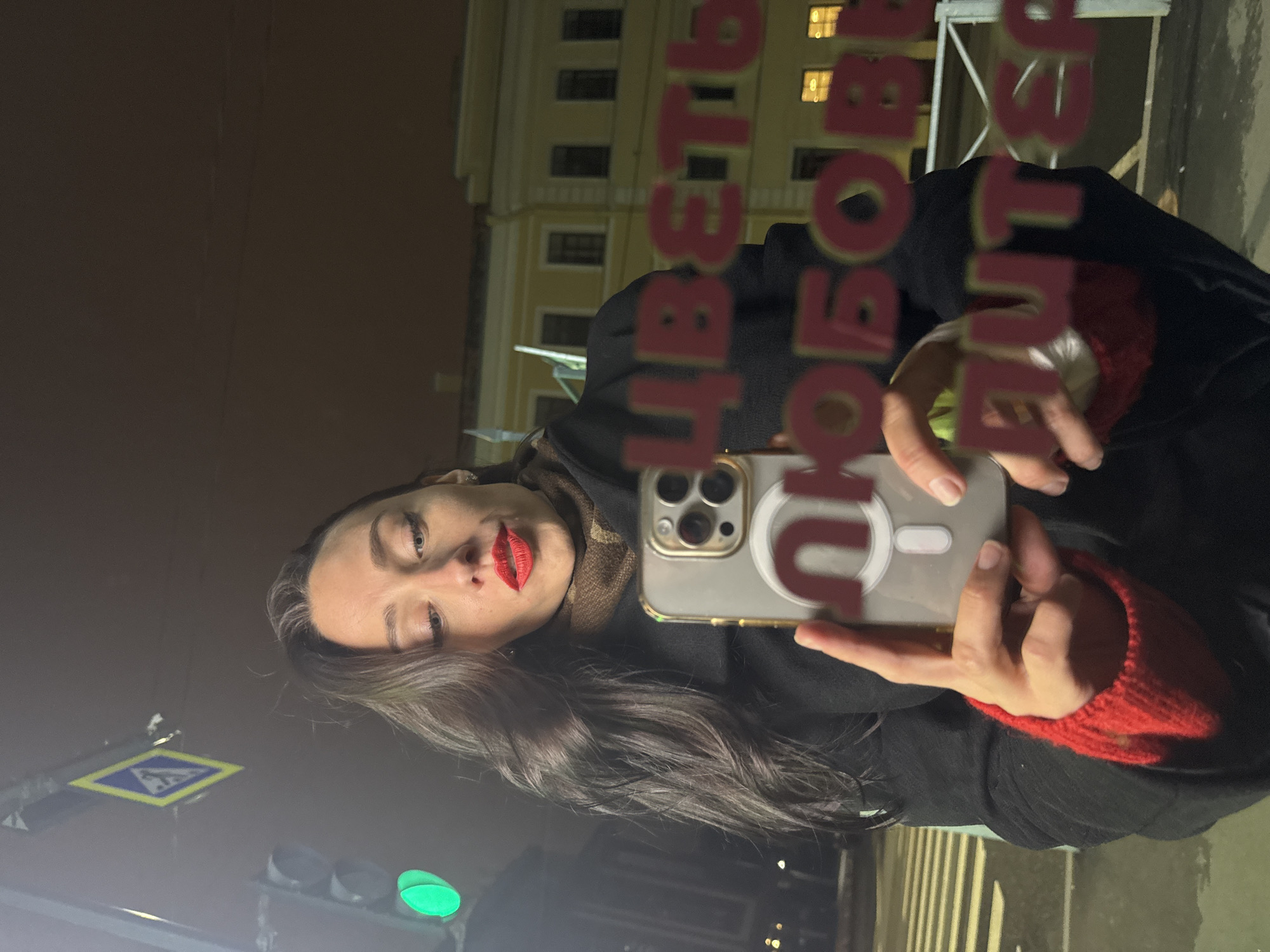 A woman with long hair and red lipstick is taking a selfie with a phone in Saint Petersburg, Russia, at night. The reflection shows a street, a building with lit windows, and a pedestrian crossing sign. The words "ЦВЕТЫ ЛЮБОВЬ И СЧАСТЬЕ" (Flowers, Love, and Happiness) are reflected in the glass.