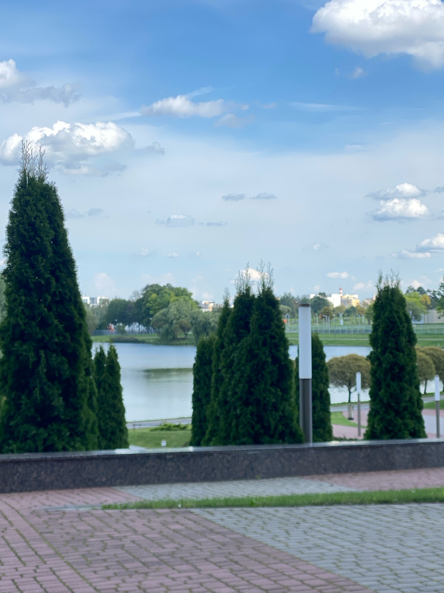 The image depicts a serene outdoor scene with a focus on a body of water, likely a lake or river, in Minsk, Belarus. In the foreground, a paved walkway composed of reddish-pink and gray interlocking stones leads the eye towards a low, dark gray stone wall. Beyond the wall, lush green grass is visible, bordered by a line of evenly spaced, tall, conical evergreen trees, characteristic of landscaping in urban parks. These trees frame a tranquil body of water, with its surface reflecting the partly cloudy sky.
In the mid-ground, across the water, a treeline of mixed deciduous and evergreen trees forms a verdant backdrop. Further in the distance, the cityscape of Minsk is subtly visible, with buildings appearing as muted shapes against the horizon. Several modern-looking structures, possibly apartment buildings or offices, can be discerned, some featuring white facades and accents of red and yellow. The sky above is a vibrant blue, dotted with fluffy white cumulus clouds, suggesting a pleasant day.
The overall impression is one of urban tranquility, where natural elements like water and trees are integrated with the cityscape, creating a pleasant environment for leisure or contemplation. The lighting suggests it is daytime, with the sun likely diffused by the clouds, creating soft shadows. No people or animals are visible in the frame, emphasizing the peaceful and unoccupied nature of the scene. The presence of paved walkways and manicured trees strongly suggests a public park or recreational area within Minsk.