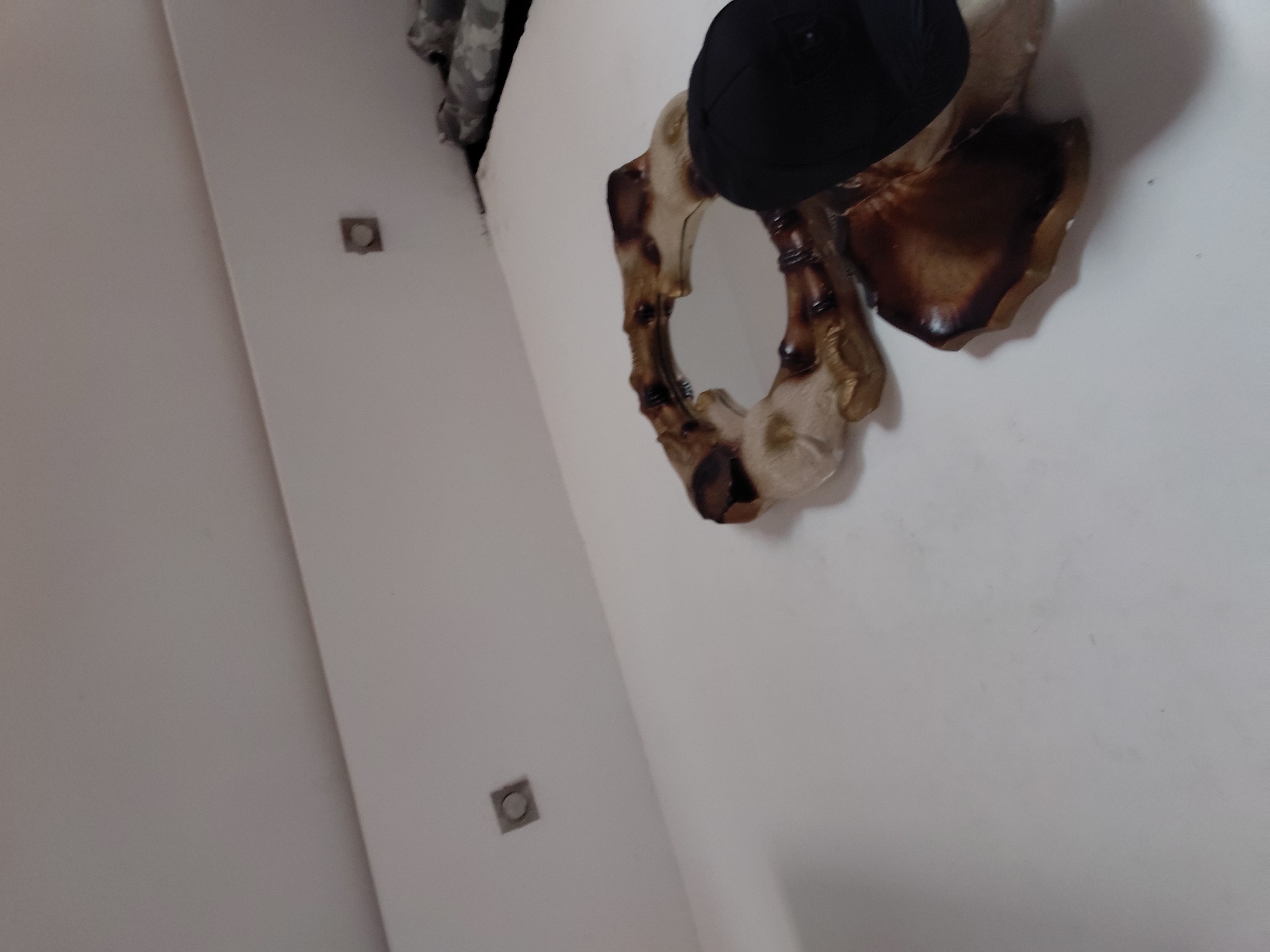 An indoor scene is captured from a tilted, upward-looking perspective, focusing on a wall decorated with a distinctive mirror and a cap. The central object is a decorative mirror, featuring an intricately carved frame that appears to be made from wood or a similar material, resembling intertwined roots or coral. The frame has a rich, varied texture and color palette, transitioning between lighter beige tones and darker, burnished browns. A black baseball cap rests on top of this mirror.
The walls of the room are a plain, clean white. Two square, recessed light fixtures with circular central elements are visible on the left wall, indicating interior lighting. In the upper left corner of the frame, a small section of a patterned fabric, possibly grey and white camouflage or a similar mottled design, is partially visible. There are no people present, no specific activity occurring, and no visible text. The time of day or external weather conditions cannot be determined from this indoor shot. The setting appears to be a modern and well-maintained room in Santa Ana, Venezuela.