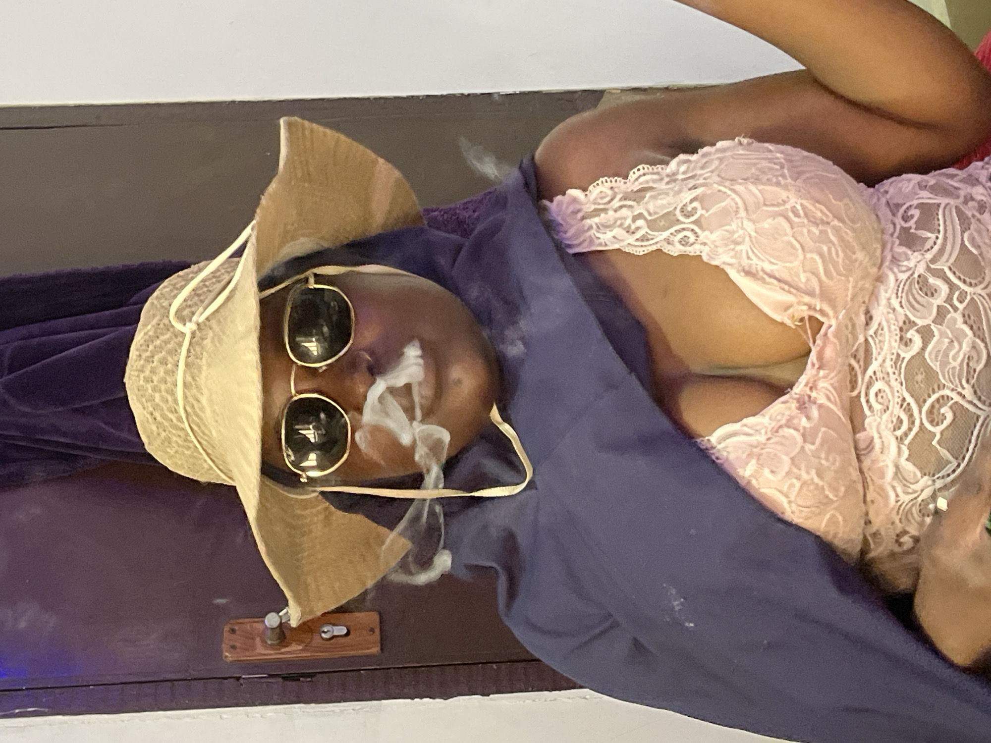 The image shows a person wearing a light pink lace top, a tan straw hat, and sunglasses. Smoke is visible in front of their face. The person is lying on what appears to be a dark blue or purple fabric. A door with a lock is visible in the background. It is impossible to confirm the location is Gwarinpa, Nigeria based solely on the image.
