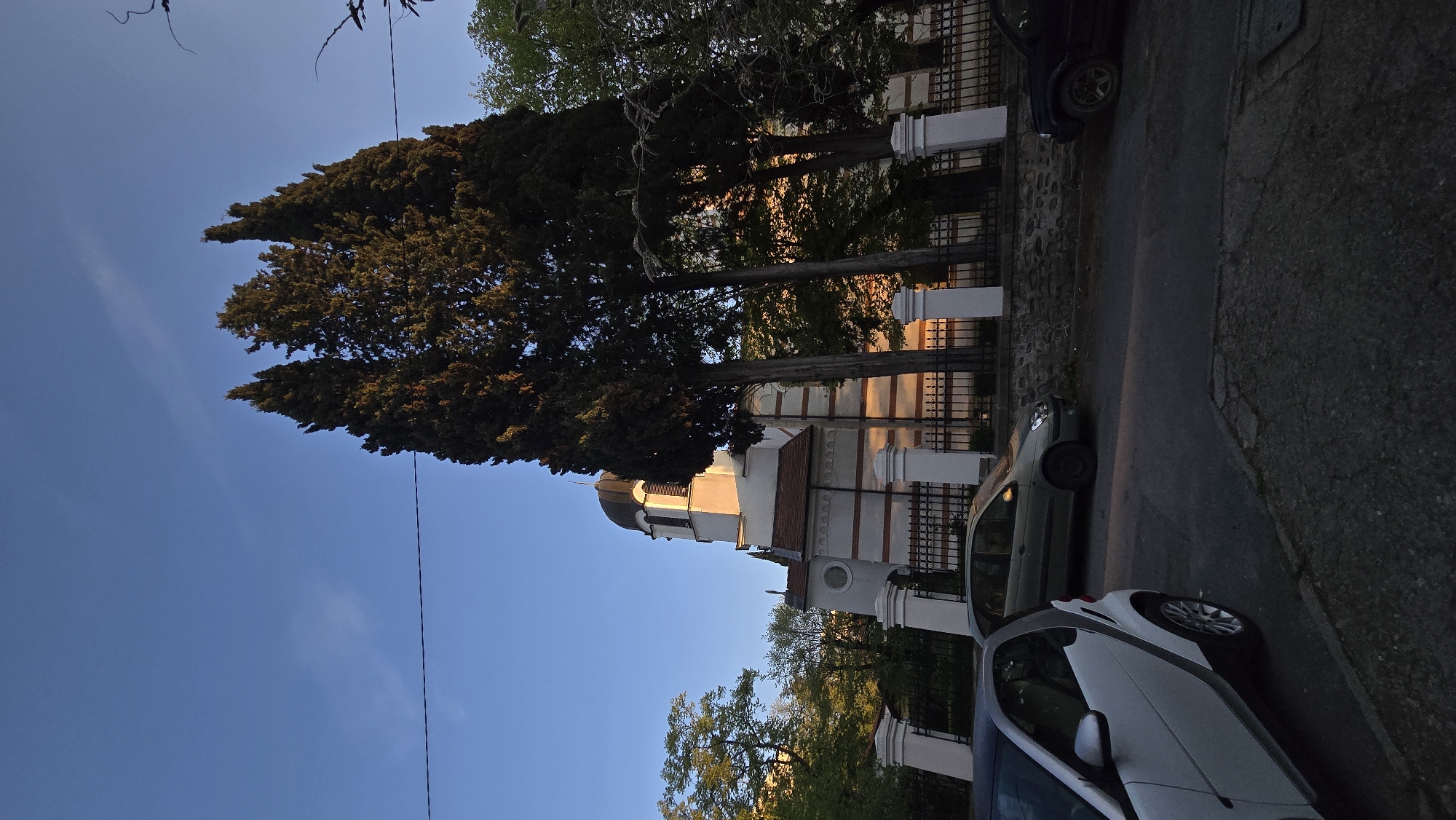Here is a description of the image:
The image shows a street scene in Stara Zagora, Bulgaria. On the left side, a building with a white and red striped facade, topped with a black dome, can be seen. Trees are planted along the side of the building. On the right, two cars are parked on the street. A black iron fence runs alongside the building and the parked cars. The sky above is a clear blue.
