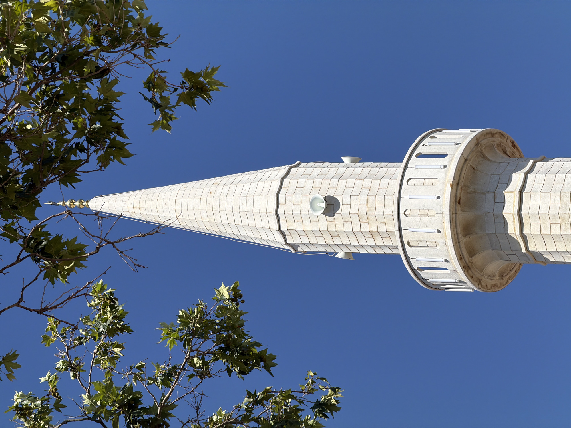 This low-angle outdoor, daytime shot shows the top portion of a minaret against a clear, deep blue sky. The minaret is constructed of light-colored stone blocks and has a conical roof topped with a gold finial. Around the middle of the visible portion of the minaret, there is a circular balcony with a white railing. Several white speakers are visible mounted on the sides of the minaret. In the upper left and top of the frame, green leaves and branches from a tree partially obscure the view, suggesting the minaret is surrounded by foliage. The scene is likely located in Ulcinj, Montenegro, a city known for its historical mosques and minarets.