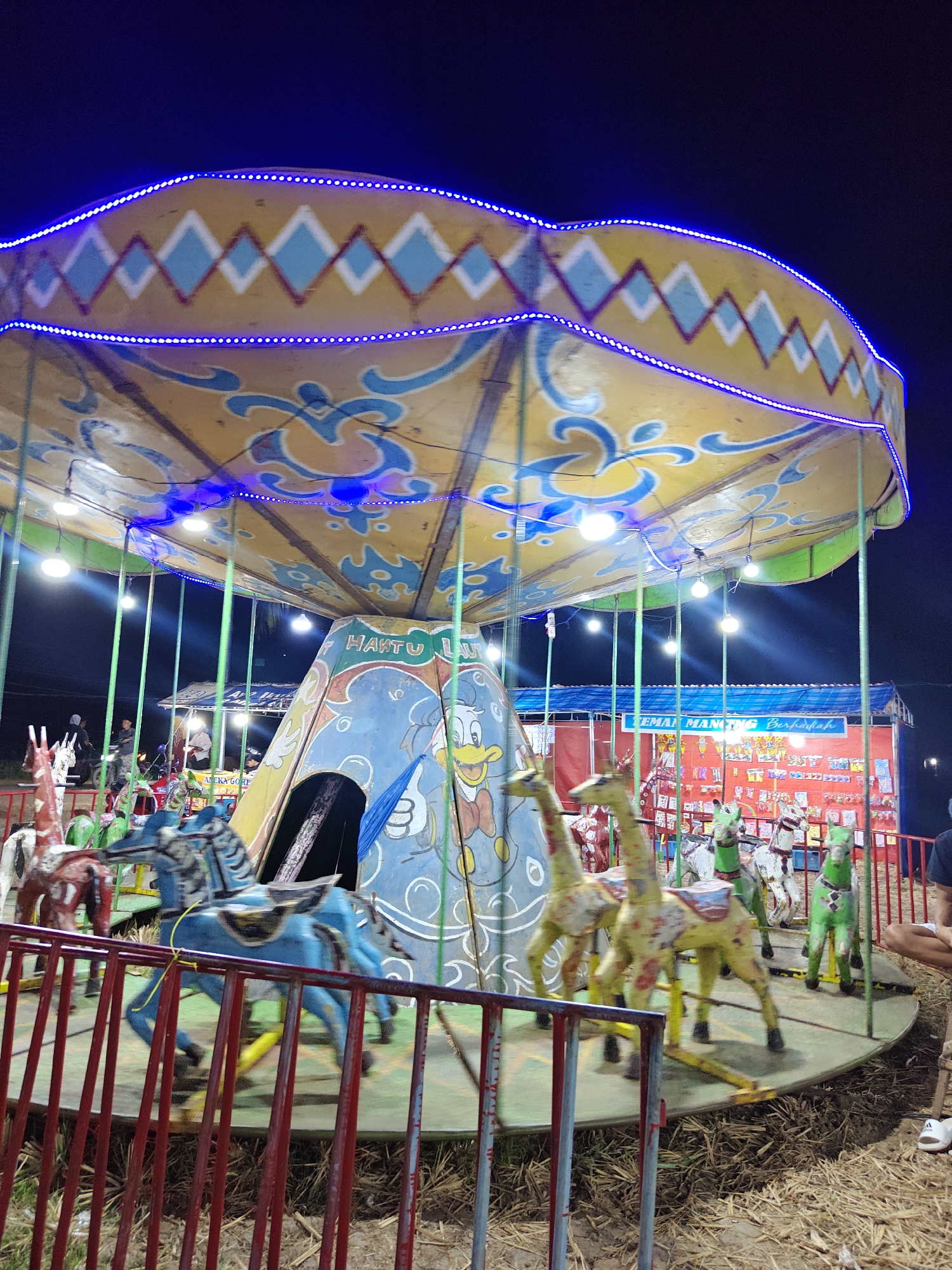 An outdoor carousel at night in Suling Wetan, Indonesia, features brightly colored, motion-blurred fiberglass animals. The carousel's top is adorned with blue lights and yellow and blue diamond patterns. A central structure on the carousel is decorated with a cartoon duck and the text "HANTU LAUT". In the background, a red and blue stall with various items on display, possibly for sale or as prizes, is illuminated. Several individuals are visible near the stall and the carousel, one standing and two seated, observing the scene. The ground is covered with straw and dried grass. The overall impression is of a lively night market or fairground attraction.