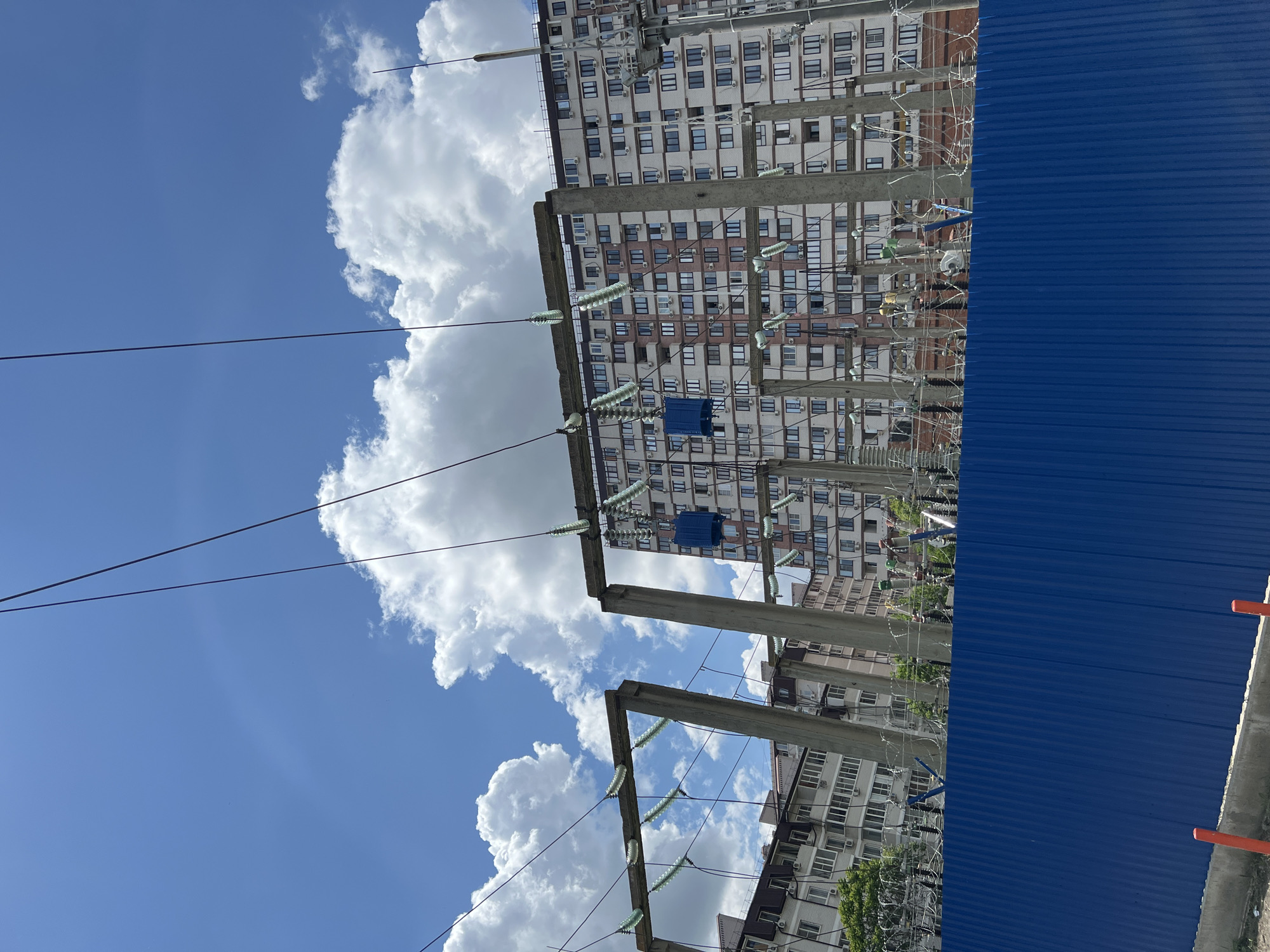 The image depicts a power substation with transmission lines and insulators against a backdrop of a residential building in Krasnodar, Russia. The substation has concrete support structures and a blue corrugated fence. The sky is blue with cumulus clouds.