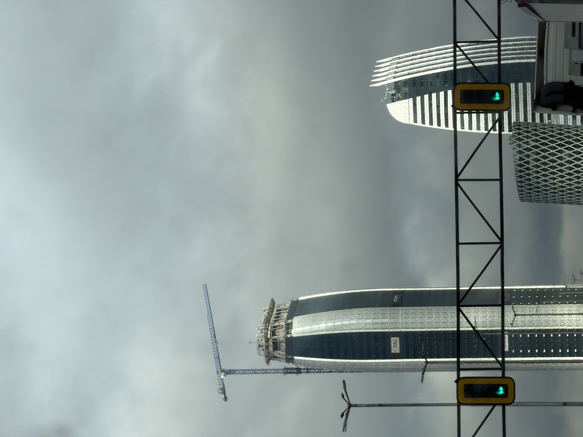 The image shows a view of buildings and infrastructure in Sofia, Bulgaria. The sky is overcast. Two modern skyscrapers are visible. The lower skyscraper is under construction with a blue crane attached. The upper skyscraper has a layered design. A traffic signal with a green arrow is in the foreground.