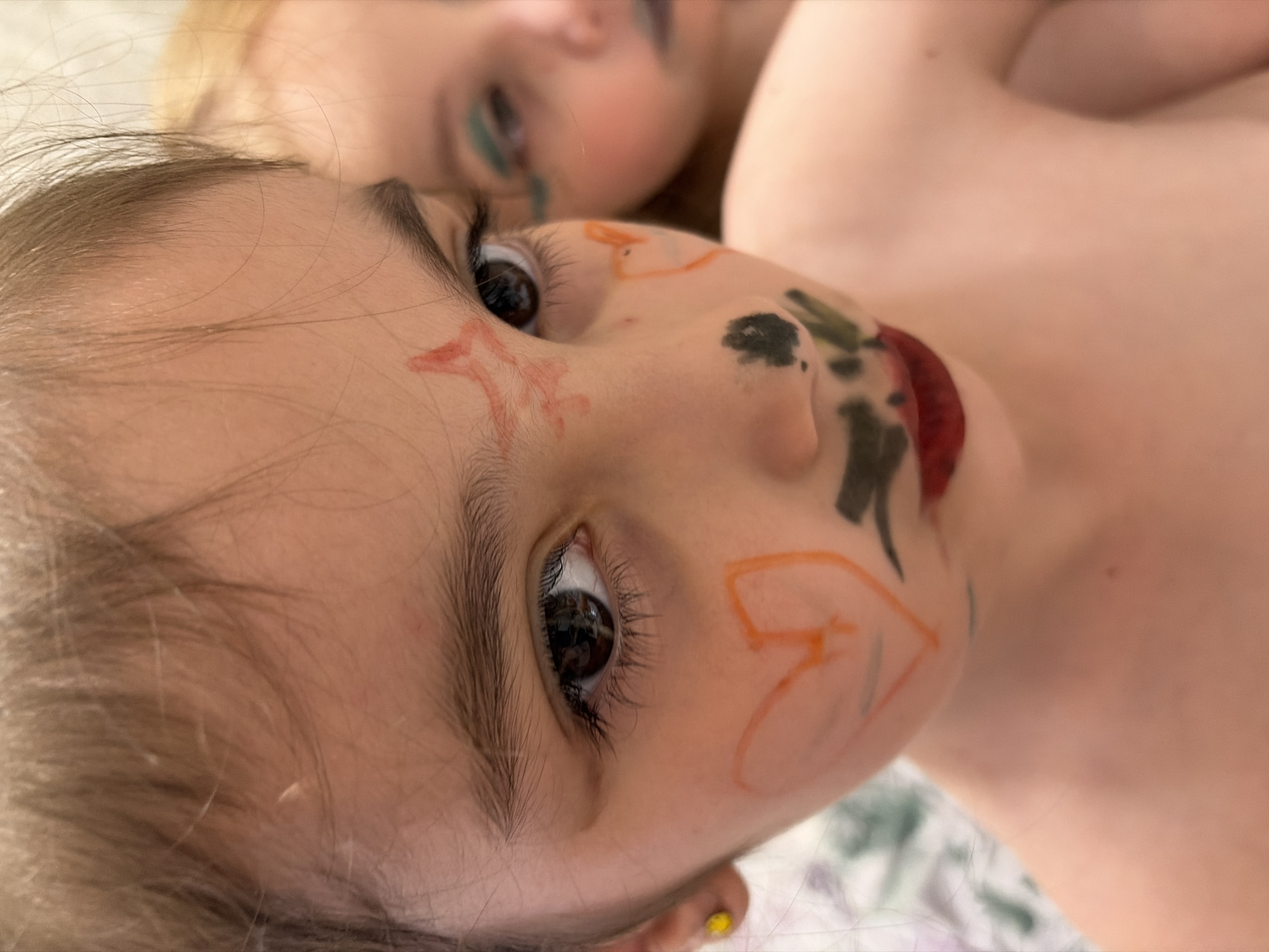 The media file is a close-up, vertically oriented photograph featuring two young children, likely siblings, with face paint. The child in the foreground, positioned on the left, has light brown hair and is looking towards the camera. Their face is adorned with orange and red markings, including a star shape on their forehead and lines and shapes on their cheek. A dark blob of paint is also visible on their cheek, and their lips have red paint. The child's right eye is clearly visible, reflecting light.
Behind and slightly above this child is another child, whose face is out of focus. This second child has blonde hair and appears to have green eyeshadow or face paint on their eyelids and under their eyes. Their mouth is slightly open.
The children are positioned close together, suggesting an intimate or playful moment. The lighting appears to be natural, possibly from a window, and the overall mood seems lighthearted and playful, given the face paint. The background is blurred but hints at a patterned surface, possibly bedding or a blanket. The image is likely taken indoors, in a home environment. No text is visible in the image. The location context provided is Tambov, Russia, but there are no specific visual cues within the image to confirm this.