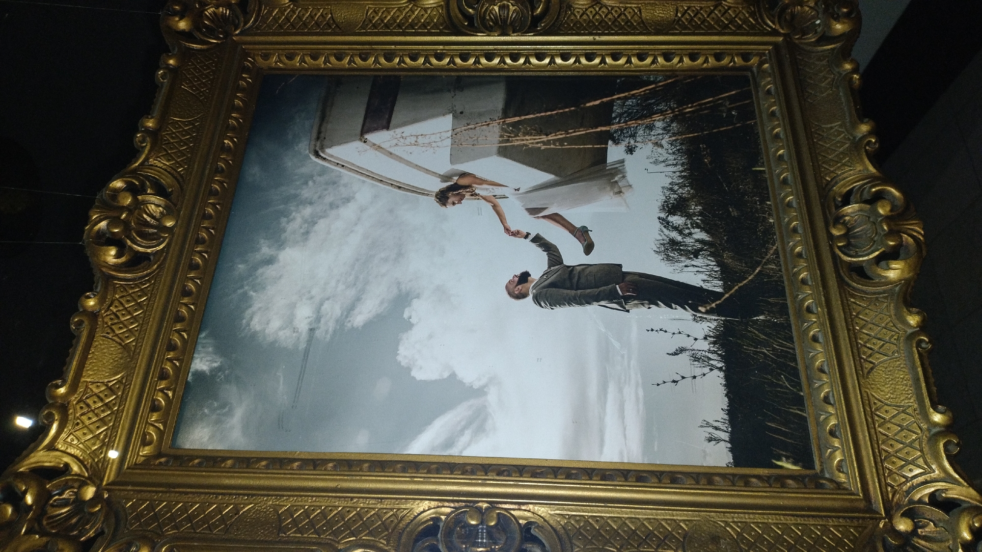 The image shows a framed photograph. The photograph itself is oriented such that the top of the frame appears at the bottom of the image, and vice versa. It depicts two people, a man and a woman, in an embrace. The man, wearing a suit, is looking up at the woman. The woman, wearing a white dress and heels, is looking down at the man and extending her hand. They appear to be dancing or in mid-air. Behind them, there is a backdrop of clouds and trees, and a structure that resembles a lighthouse or a tower. The scene suggests a romantic or whimsical moment. The photograph is enclosed in an ornate, golden frame with intricate carvings. The lighting in the scene within the photograph appears to be natural, possibly daytime, with the sky being overcast. The location context is Perea, Greece, but there are no specific visual cues in the photograph to confirm this. There is no visible text in the image.
