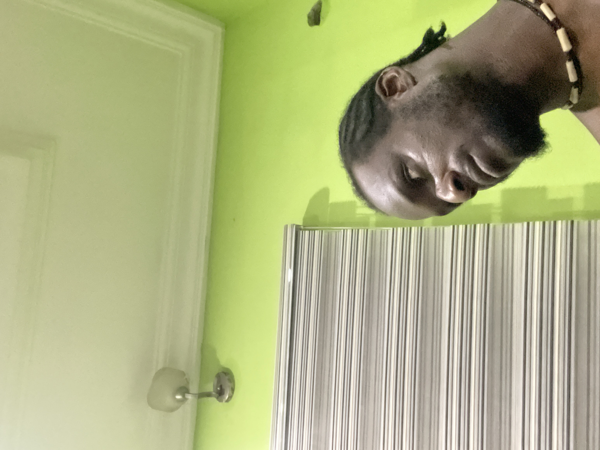 A medium shot shows a man’s face, neck, and a portion of his shoulder. He has dark skin, braids, and a short beard. He is wearing a beaded necklace. Behind him is a green wall and part of a vertically striped blind or screen. In the background, on the left, is part of a white door, including the doorknob and frame. The location is Abuja, Nigeria.