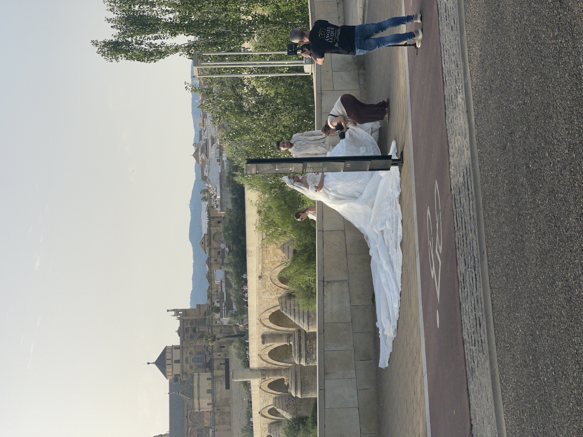 In Córdoba, Spain, a wedding photoshoot is taking place on a pedestrian bridge overlooking the Guadalquivir River. A bride in a voluminous white wedding dress with a long train is posing, while a groom in a light-colored suit stands beside her. A photographer, wearing a black t-shirt with white lettering that reads "ANGEL LUQUE FOTÓGRAFO," is capturing the moment. Another person is kneeling and adjusting the bride's dress, seemingly assisting the photographer. In the background, the historic Mezquita-Cathedral of Córdoba is visible, along with other city buildings and lush trees lining the riverbank. The scene is set against a clear sky, suggesting a pleasant day.