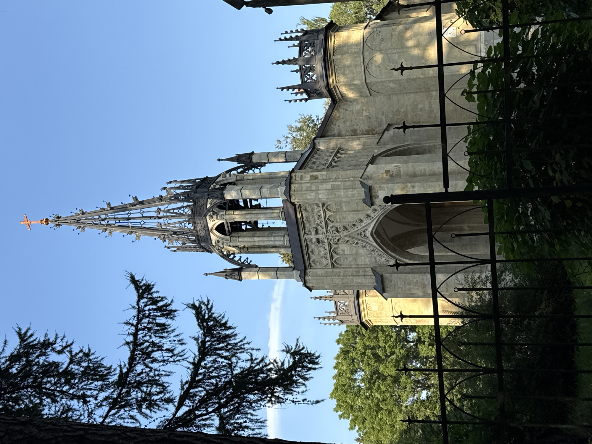 A Gothic Revival chapel, likely a mausoleum, stands against a clear blue sky, partially obscured by the dark silhouette of pine branches in the foreground. The chapel is constructed from light-colored stone, featuring intricate architectural details such as pointed arches, decorative tracery, and a tall, spired tower topped with a golden cross. A black metal fence runs across the lower portion of the frame, in front of lush green foliage. Trees with vibrant green leaves surround the structure. The scene appears to be a serene, historical site, possibly located within a park or cemetery. This chapel is identifiable as the Mausoleum of Prince Alexander Menshikov, located in the Ohta Park (formerly part of the estate of Empress Maria Fyodorovna) in Pargolovo, Russia.