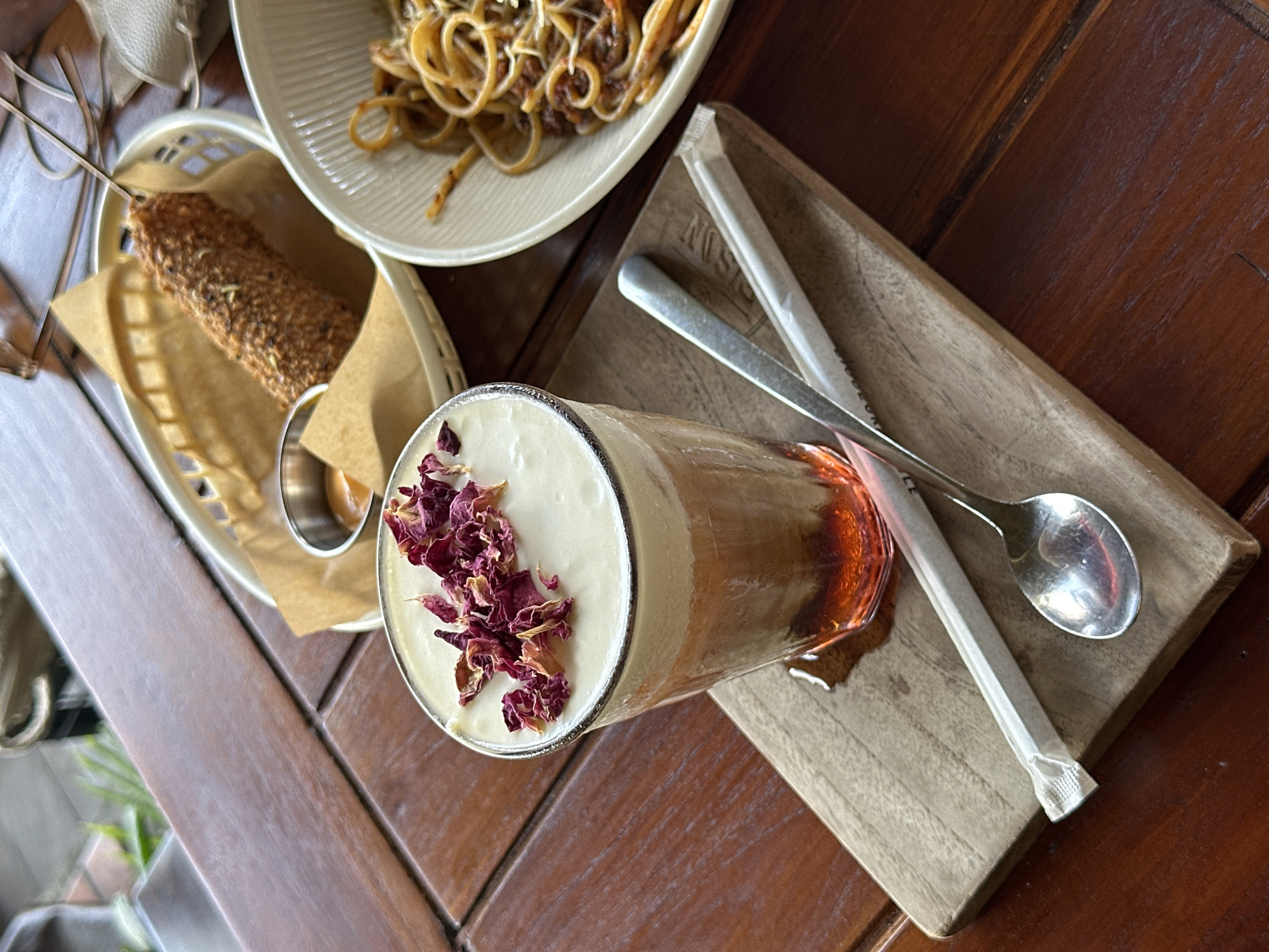 This image shows a tabletop scene with various food and drink items in Ubud District, Indonesia. There is a cylindrical glass of layered beverage topped with dried rose petals, positioned on a small wooden cutting board along with a spoon and a wrapped straw. Next to it, there's a basket containing a skewered, breaded food item, possibly a snack or appetizer. Partially visible in the background is a plate of pasta. The table is made of dark wood.