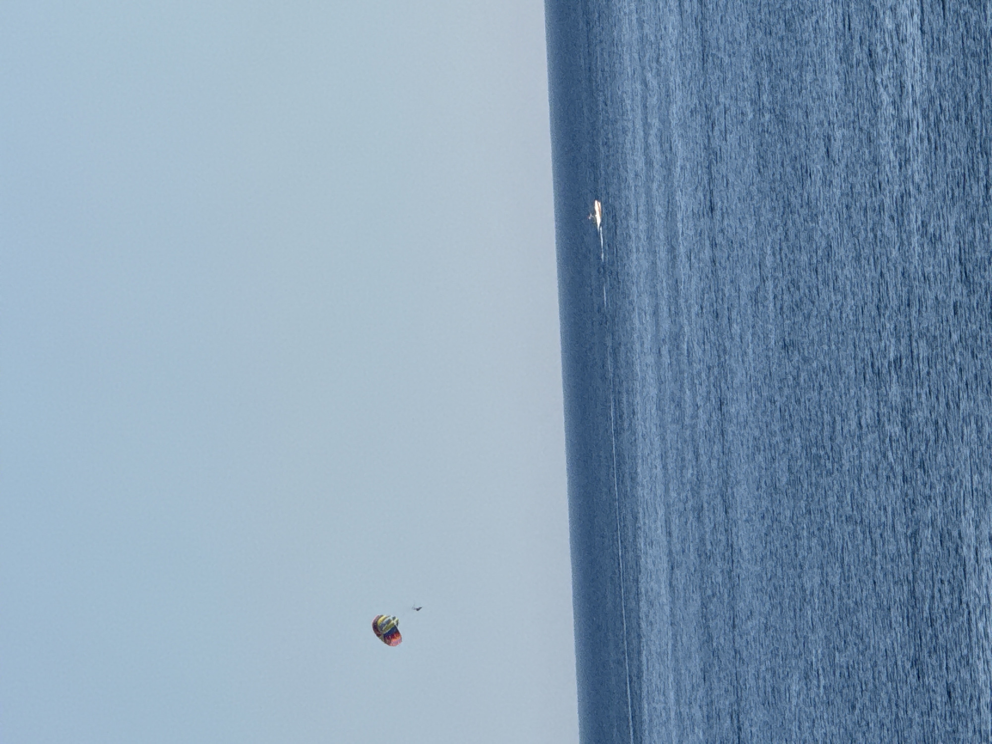 A wide shot captures a serene seascape under a clear, pale blue sky. The image is divided horizontally, with the upper half dominated by the sky and the lower half by the deep blue ocean. The ocean surface is textured with subtle ripples and reflects the sky's light. In the distance, a small, white motorboat navigates the water, leaving a faint white wake behind it. Above the horizon, a colorful parachute, red, yellow, green, and blue, floats in the sky, tethered to an unseen person or object. The composition suggests a peaceful day at sea, likely in a coastal area such as Kamenovo, Montenegro, known for its scenic beaches and water activities.