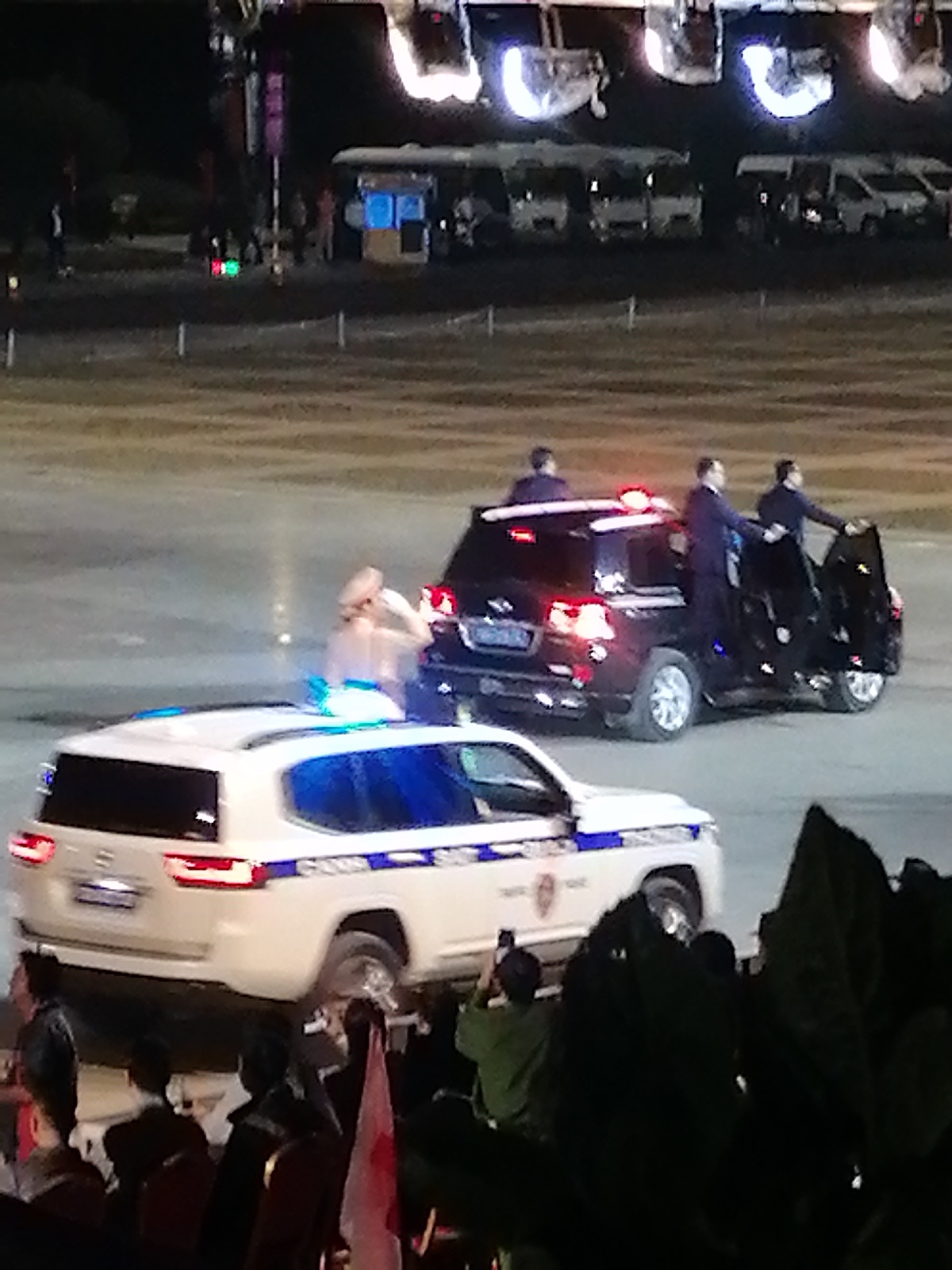 In Da Lat, Vietnam, a motorcade is captured at night, featuring two black SUVs and a white police vehicle. The leading SUV's rear lights are illuminated, and two individuals in dark suits are seen exiting the vehicle. One man is in the process of closing the passenger door while the other appears to be shaking hands. The second SUV is partially visible with its rear lights on, and a person in a light-colored garment is standing near its open passenger door. In the foreground, the white police car has its emergency lights flashing. A crowd of spectators is gathered, some holding flags and others taking photos. The scene is set on an open, paved area, possibly a street or a designated event space, with blurred background elements suggesting structures and lights.