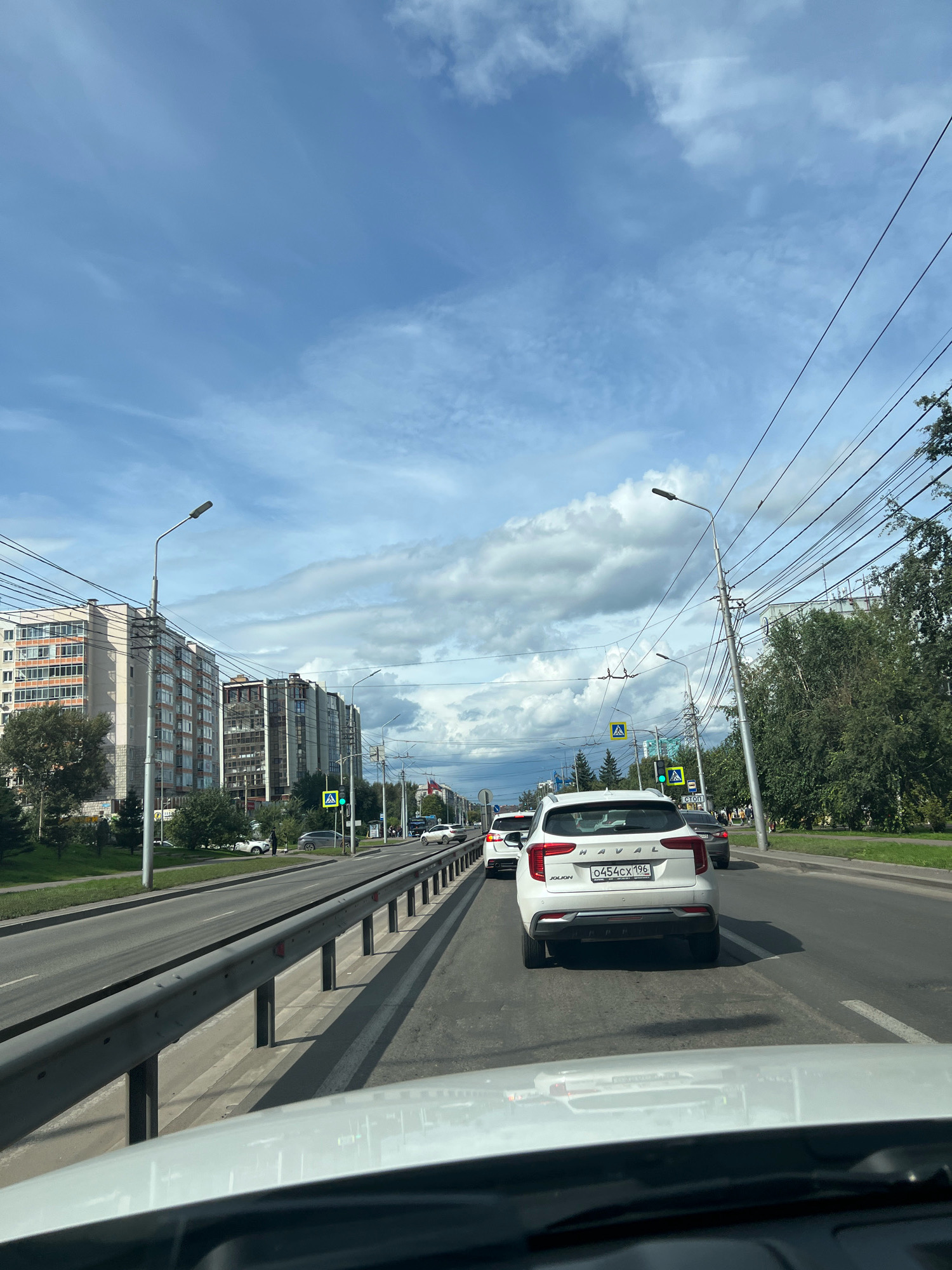 The image depicts a view from a vehicle traveling on a multilane road in the city of Krasnoyarsk, Russia, under a partly cloudy sky. The road is lined with trees and buildings on the left side, including residential apartment buildings with balconies and windows. On the right side, there are more trees and a sidewalk. Electrical wires are visible strung across the scene, supported by poles. Traffic lights and streetlights are also present. Several cars are on the road, indicating moderate traffic. The vehicle from which the photo is taken is in the foreground, with the hood and windshield visible. One car in front, a white Haval JOLION, has a license plate that reads "O 454 CX 196". Pedestrians are visible on the sidewalk to the right.