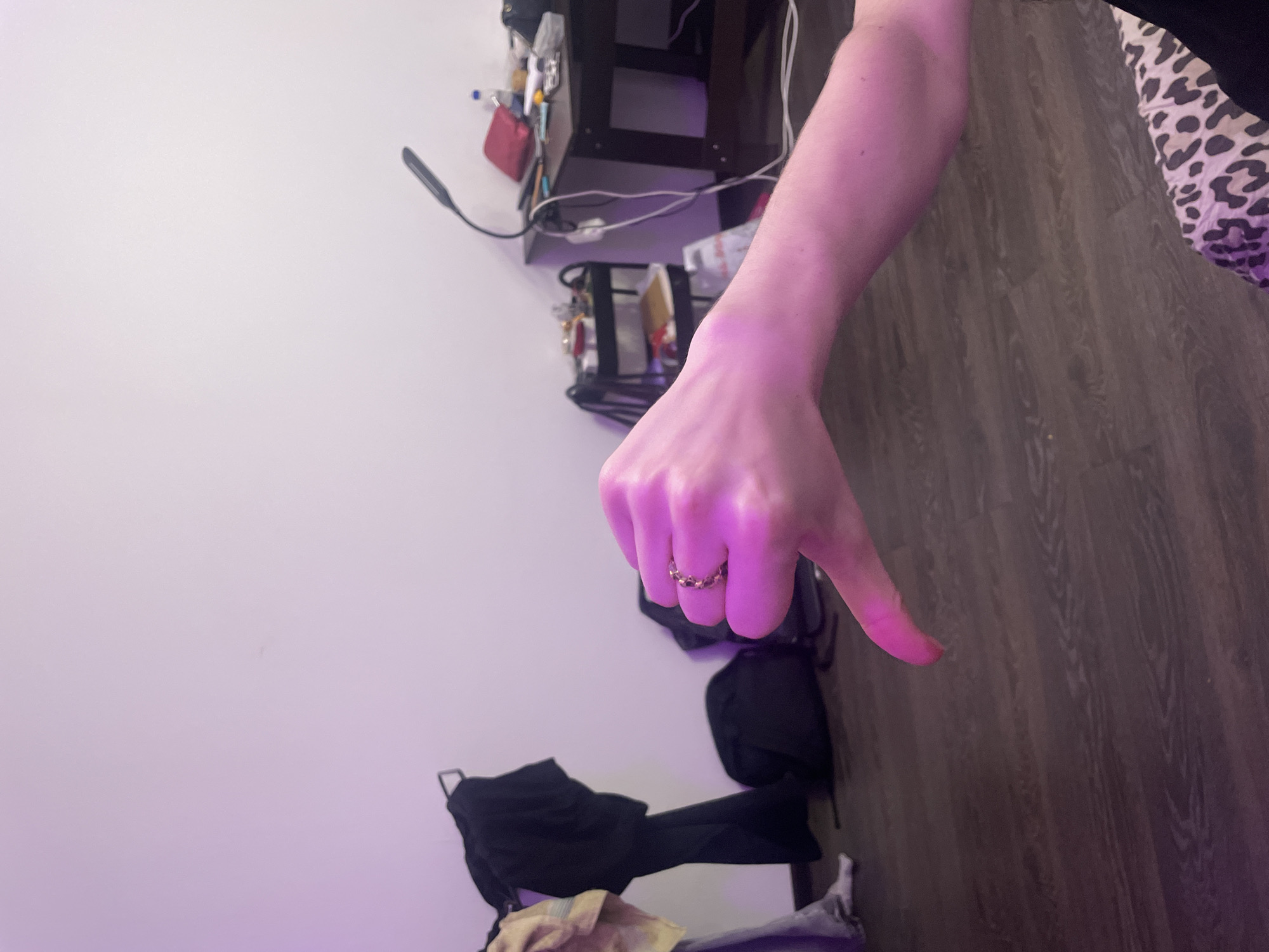 In an indoor setting, likely a bedroom or dorm room, a person's right hand is prominently displayed in the foreground, giving a thumbs-down gesture. The hand is illuminated by purple lighting, casting a pinkish hue over the skin. A ring with dark, possibly red, stones is visible on the ring finger. The background reveals a dimly lit room with wooden flooring. To the left, several items of clothing are draped over a rack, including a black jacket and a lighter-colored garment. A black backpack rests on the floor behind the clothing. Further into the background, on the right side of the image, a desk with various items like a red pouch, a bottle, and other unidentifiable objects is visible. A flexible desk lamp is attached to the desk. A small rolling cart with shelves also stands near the desk. The overall environment suggests a casual, lived-in space. There is no information in the image to suggest the location is the city of Sinyavino, Russia.