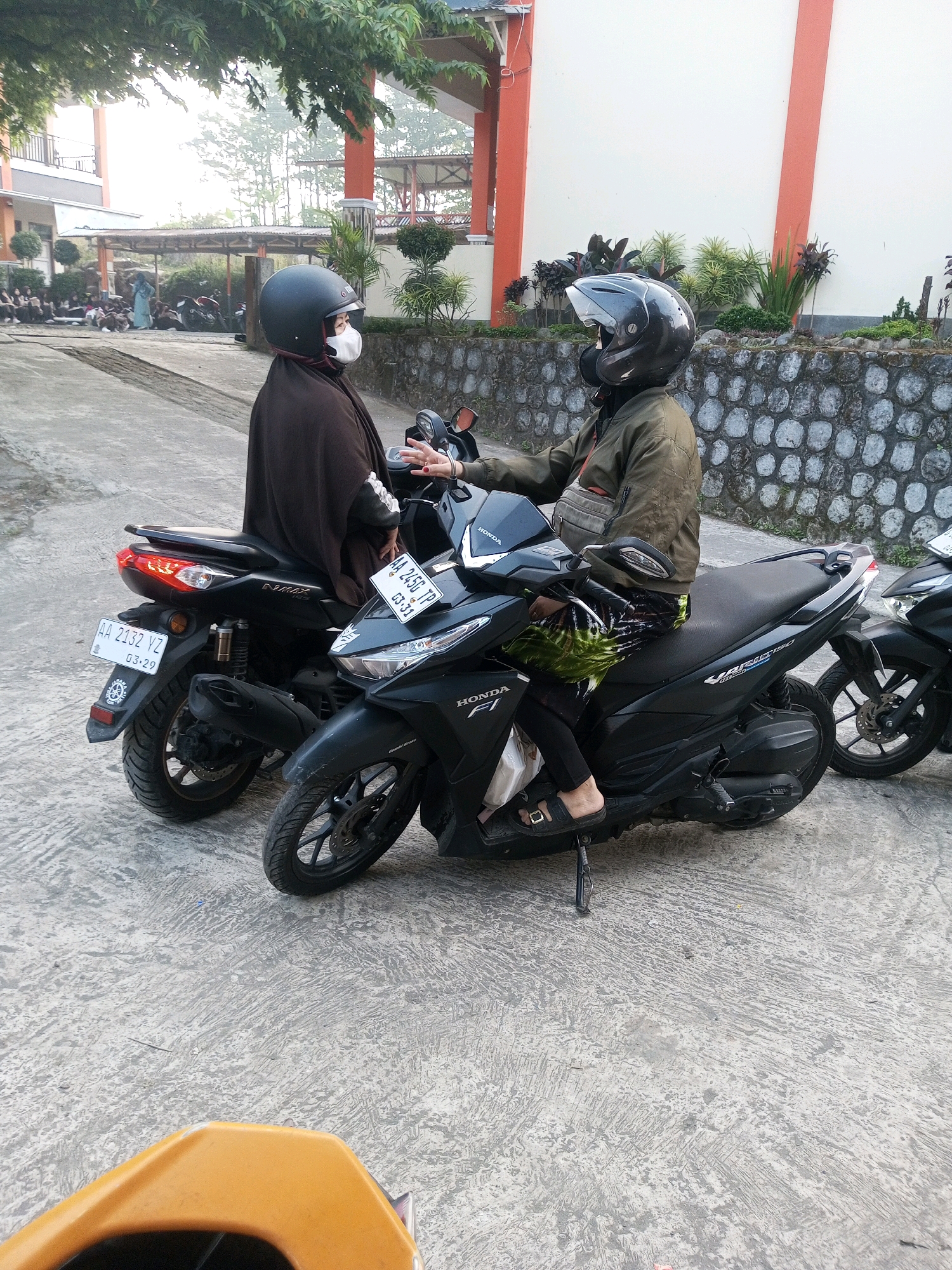 Two individuals wearing helmets are seated on motorcycles parked on a paved surface. The motorcycle on the right is a black Honda, and the motorcycle on the left is also black, identified by its license plate as "AA 2132 YZ". The individual on the left is wearing a brown head covering and a white face mask. The individual on the right is wearing a green jacket, black pants, and sandals. The background shows a building with orange trim, a stone wall, and trees. The scene appears to be an outdoor area in Krasak, Indonesia, possibly near a school or public facility, given the presence of other people in the background who are seated on the ground. The lighting suggests it is daytime, possibly early morning or late afternoon due to the soft light.