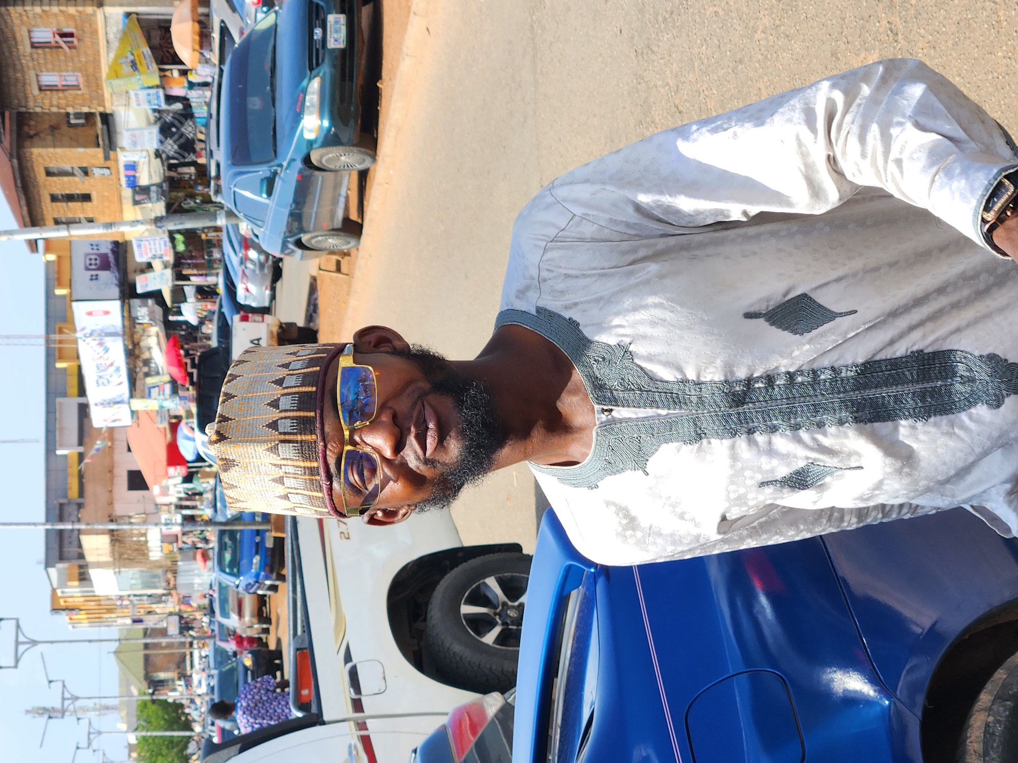 The image captures a vibrant street scene under bright daylight, indicative of an urban commercial area, consistent with the context of Jos, Nigeria. The main subject is a man positioned in the foreground, posing next to a blue car. He sports a dark beard and mustache, and wears reflective yellow-framed sunglasses. His head is adorned with a traditional patterned cap, likely a kufi, and he is dressed in a light-colored traditional tunic, possibly a boubou or agbada, featuring intricate grey embroidery on the chest and collar. A wristwatch is visible on his right wrist. The man gazes directly at the camera with a relaxed, slightly smiling expression.
Behind the man and the blue car, the street stretches into the background, revealing a busy thoroughfare. A white SUV or van is parked directly behind the blue car, with "2.7" visible on its side. Further down the road, several other vehicles are present, including a dark blue sedan with a partially visible license plate displaying "CM-6XX". The street is flanked by multi-story buildings constructed of brick or stone, some with visible windows. Evidence of market activity is scattered along the roadside, including various signs, merchandise, and a red umbrella, suggesting small shops or stalls. Utility poles and overhead cables line the left side of the street. The clear sky and distinct shadows indicate a sunny day, likely during midday or early afternoon. The overall scene portrays a bustling, active urban environment.