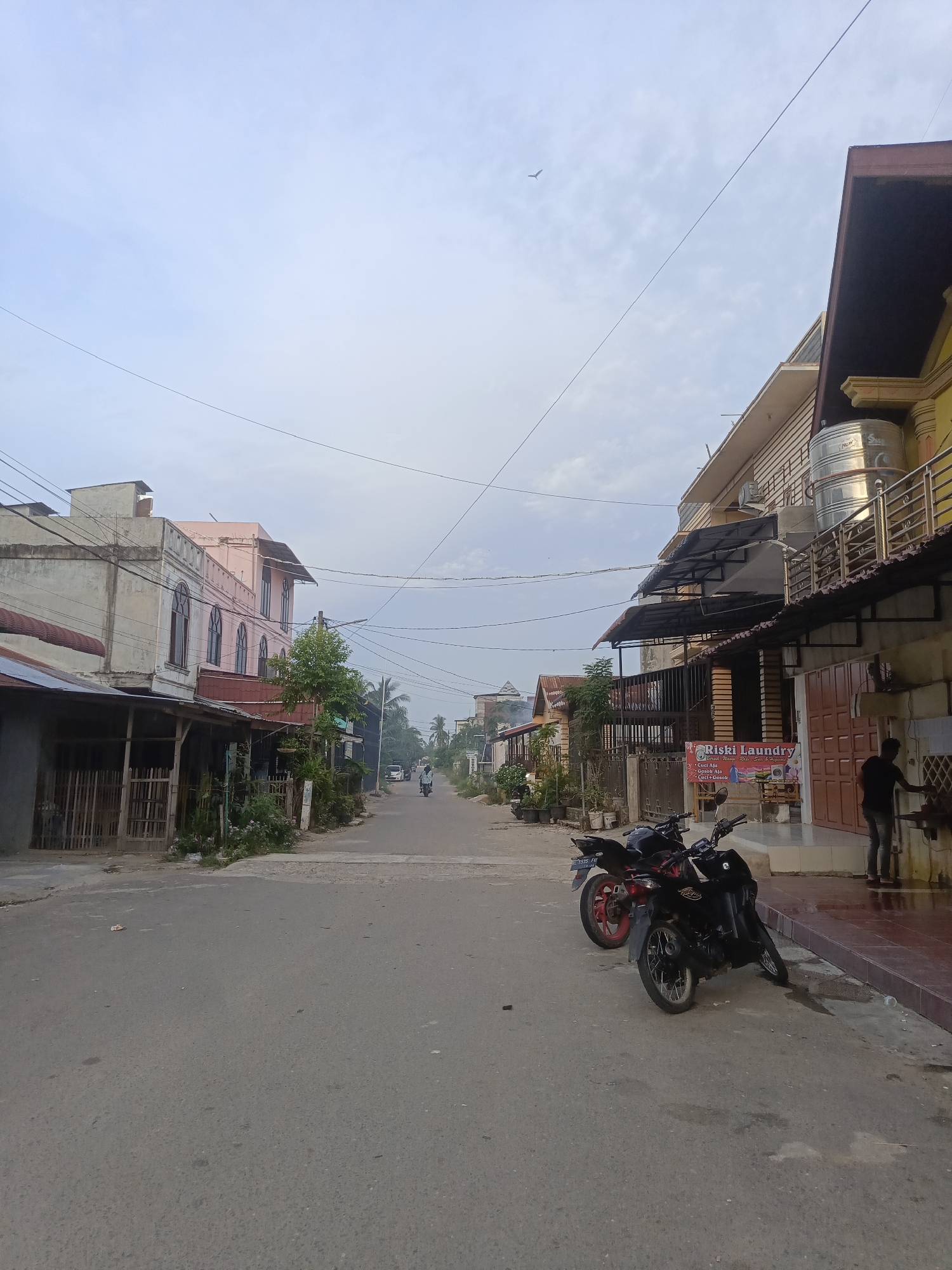 The image depicts a street scene in Lhokseumawe, Indonesia, likely in the early morning given the soft light. The street is paved and appears to be a main thoroughfare lined with buildings on both sides. On the left, there are low-rise buildings with some exhibiting a rustic, wooden facade with a corrugated metal roof, suggesting shops or residences. One building stands out with a pink, multi-story facade featuring arched windows. On the right, a more modern, multi-story building is visible, painted yellow, with balconies and a large stainless steel water tank on the roof. A laundry business, "Riski Laundry," is advertised with a banner on the ground floor. Several motorcycles are parked along the right side of the street. A single person, dressed in dark clothing, is seen standing near the laundry establishment, possibly attending to something outside. Further down the street, a person is riding a motorcycle in the distance, and a few other vehicles are scattered along the road. The sky is overcast with a pale blue hue, and utility wires crisscross overhead. Vegetation, including trees and potted plants, can be seen interspersed between the buildings. The overall impression is of a quiet, developing urban environment.