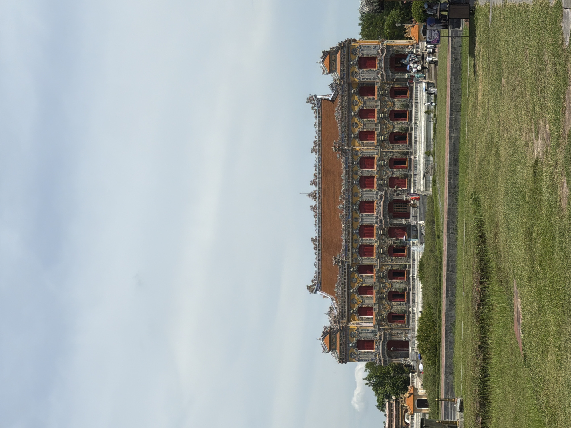 The image shows the Imperial City, also known as the Citadel, in Phú Xuân Ward, Vietnam. The Citadel is a UNESCO World Heritage Site and a popular tourist attraction. The building in the foreground is the Royal Theater, also known as the Duyet Thi Duong Theater. It is a two-story building with a red tile roof and yellow walls. The building is decorated with ornate carvings and intricate details. In front of the building is a large lawn with a few trees. In the background, there are more buildings and trees. There are also a few people in the image, most of whom are tourists. The sky is cloudy and the weather is overcast.