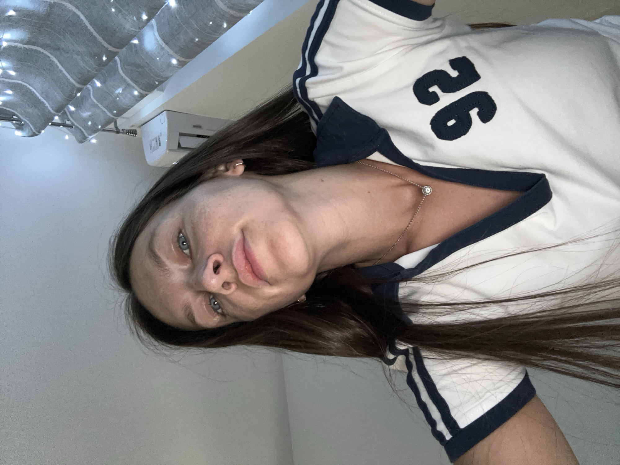 In a close-up, low-angle shot taken in a room, a woman with long, dark hair and blue eyes looks directly at the camera. She is wearing a white polo shirt with navy blue trim on the collar, sleeves, and a large "92" applique on the right side of her chest. A delicate silver necklace with a circular pendant is visible around her neck. The background shows a grey wall, a white air conditioning unit, and a window covered by striped blinds with string lights draped across them. The scene suggests an indoor setting, possibly a bedroom or living area, in Donji Pobori, Montenegro.