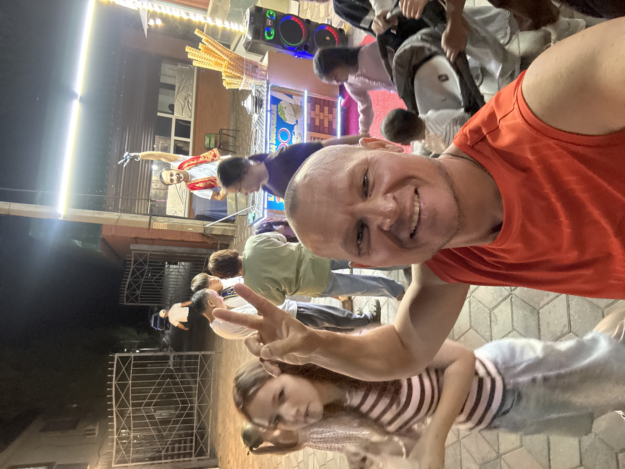 The image is a selfie taken at night, likely in an outdoor urban setting in the city of Unknown, Kazakhstan. A man in an orange t-shirt is in the foreground, smiling and holding up two fingers in a peace sign. Behind him, several other people are visible. To the left, a young girl with long brown hair wearing a striped shirt is looking towards the camera. Further back, a group of people, including men and women, are standing near an ice cream stand. The ice cream stand has a colorful display with cones stacked high and illuminated by bright lights. A man in traditional attire is standing behind the counter, holding a sign. The background shows buildings and a street. The overall atmosphere appears festive and casual.