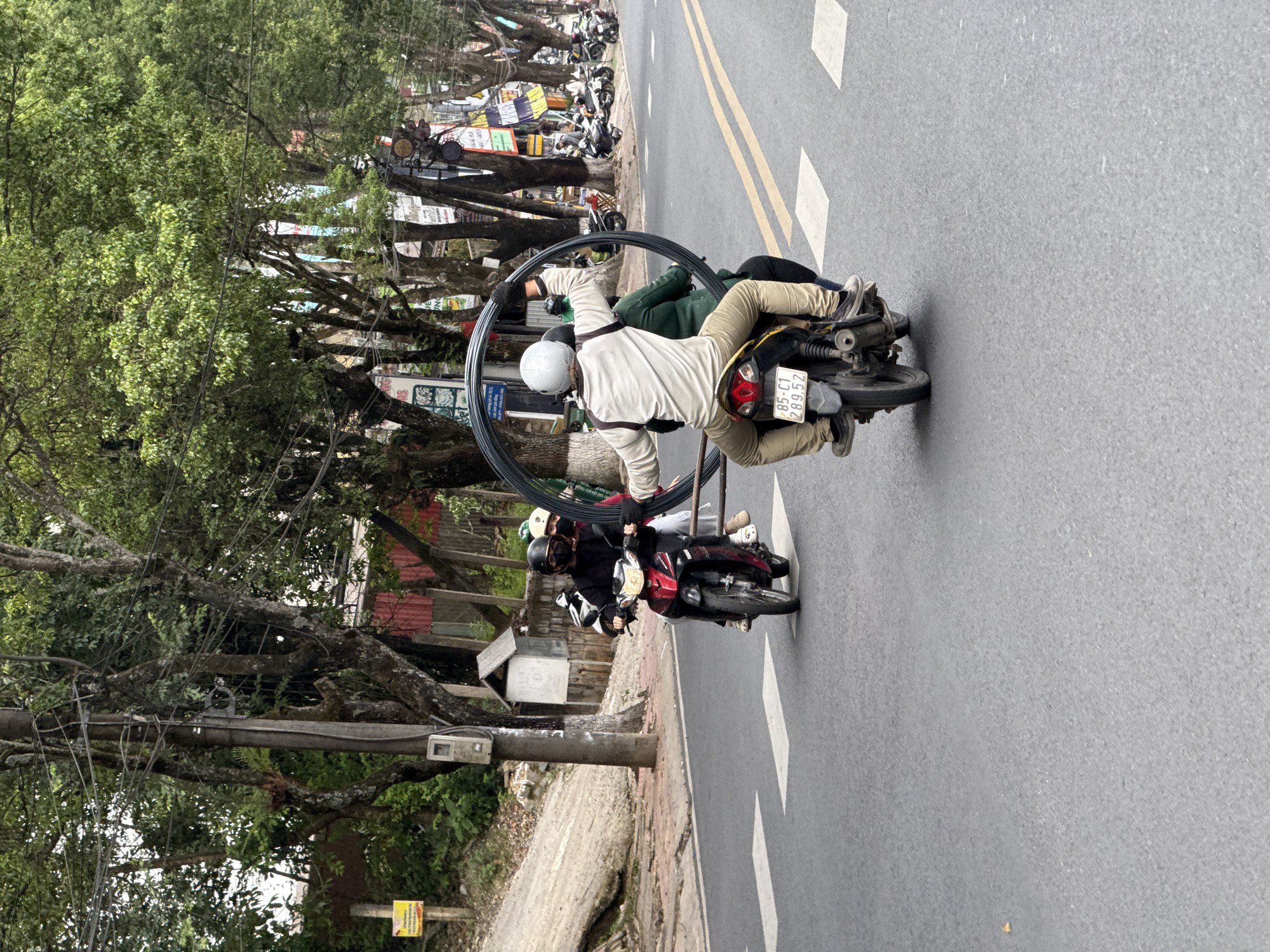 The image shows a street scene in Lâm Viên - Đà Lạt, Vietnam. A person wearing a white helmet and light-colored jacket is riding a motorcycle while holding a large circular metal object (possibly a wheel). A second person is seated behind the driver. The motorcycle has a license plate that reads "85-C1 289.52." Another motorcycle with two helmeted riders is visible behind the first motorcycle. The road has yellow dividing lines and white rectangular lane markings. The scene is bordered by trees and utility poles, and buildings and parked motorbikes can be seen further back.