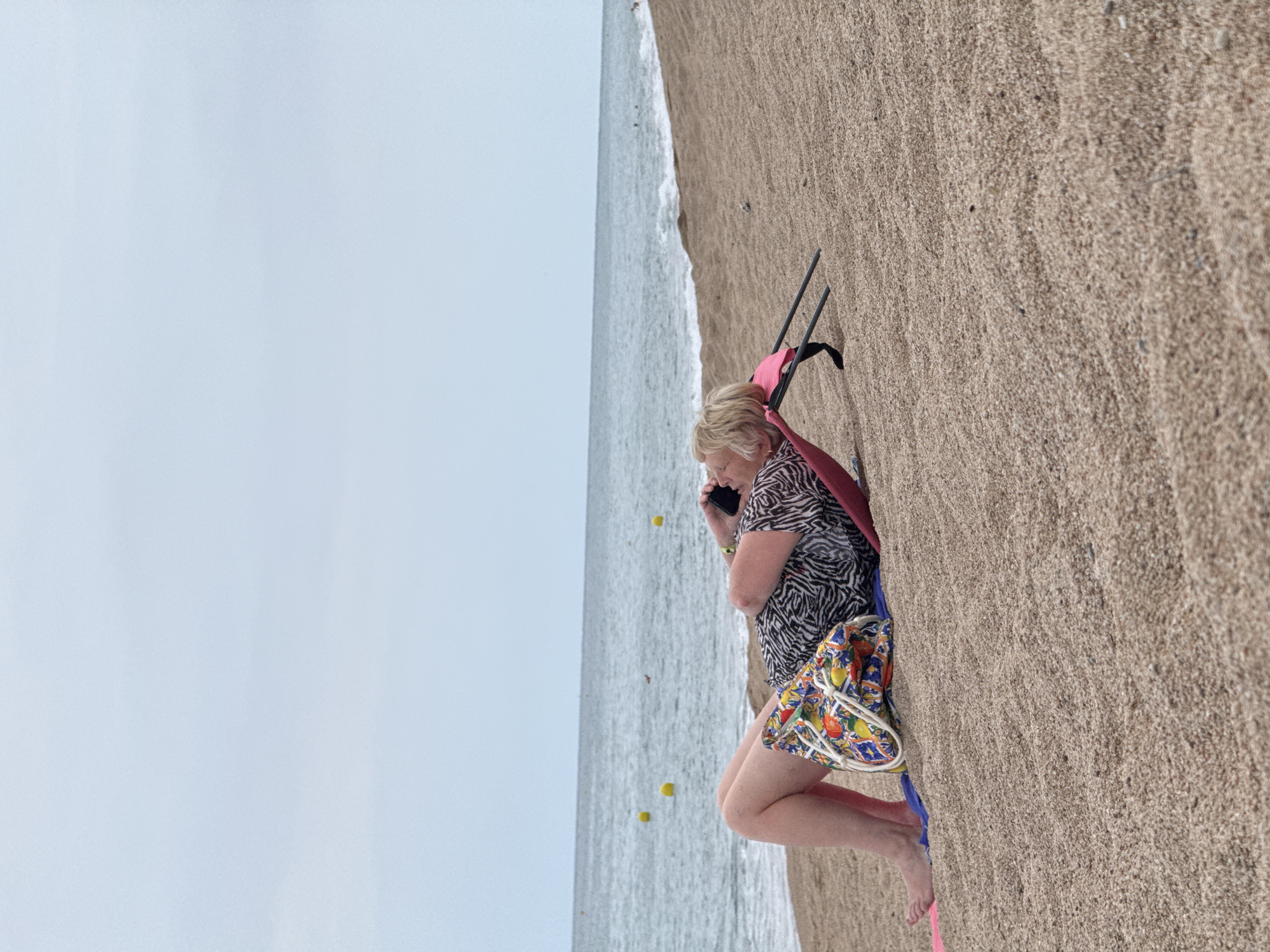 A woman is lying on a beach chair on a sandy beach, talking on her mobile phone. She is wearing a black and white patterned shirt and colorful shorts. A beach bag with a fruit pattern sits beside her. The sea is in the background, with a few yellow buoys visible in the water. The sky is overcast. The location is Blanes, Spain.