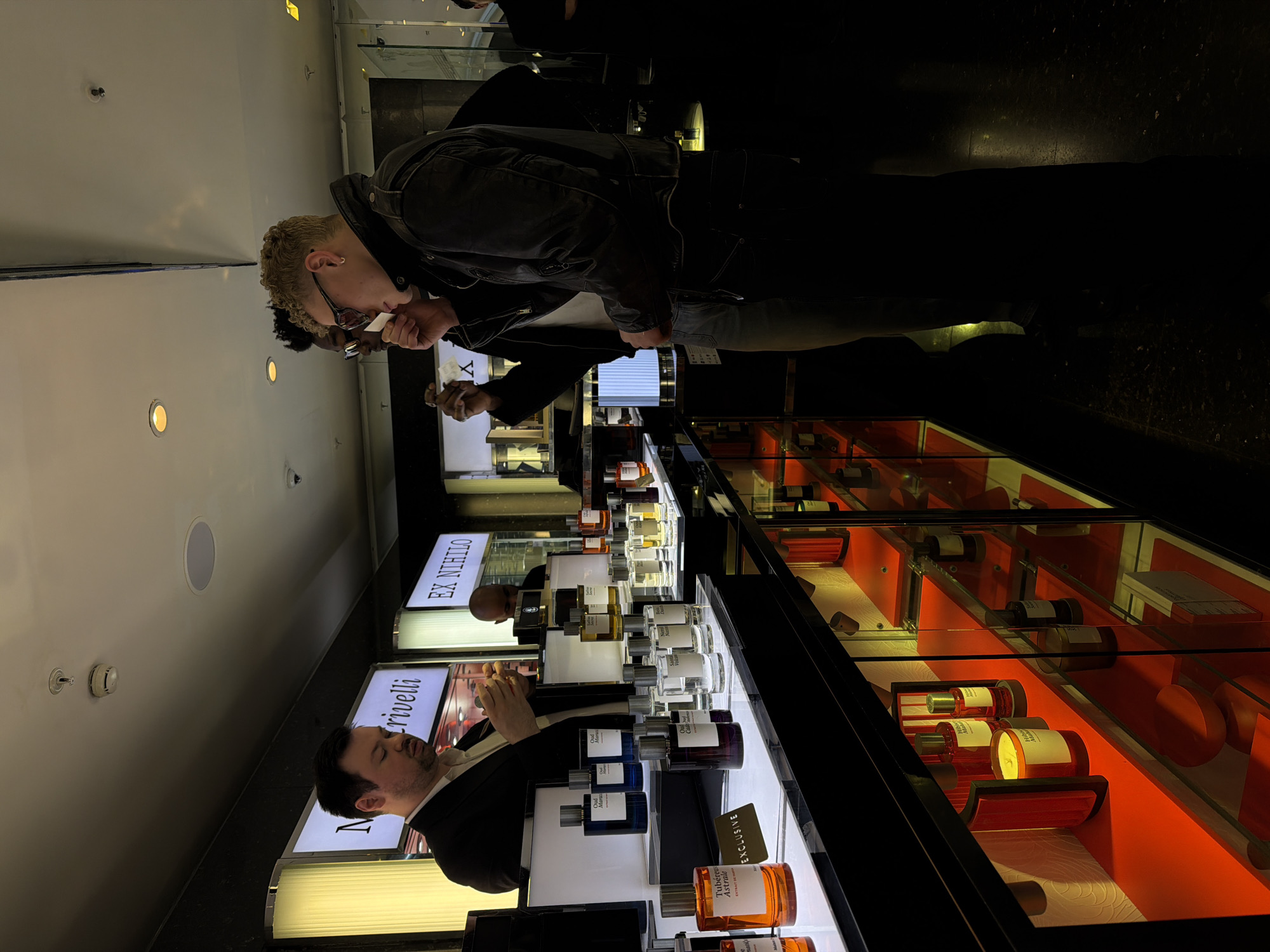 This image captures a luxury fragrance retail area within a high-end department store in London, United Kingdom. The environment is characterized by dim general lighting, contrasting with bright spotlights illuminating product displays.
On the left, a male staff member, dressed in a dark suit jacket over a light shirt, is seen from the chest up, holding a small object, possibly a perfume bottle or tester strip, near his face. To the right, two male customers are engaged in sampling fragrances. The customer in the foreground, with short blonde curly hair, sunglasses, and a dark leather jacket, holds a white blotter strip to his nose. Behind him, another male customer with dark skin and glasses is also smelling a blotter strip.
The central display counter features multiple perfume bottles of various designs, prominently arranged on lit platforms. Visible brand signage includes "M. Crivelli" (partially visible) and "EX NIHILO" (fully visible on a backlit sign). Specific fragrance bottles display labels such as "Tubereuse Astrale Extrait de Parfum" (next to a golden "EXCLUSIVE" sign), "Oud Maracuja," "Neroli Nautica," and "Santal Volcanique." Below the main counter, glass display cases with red-lit interiors showcase additional products, including candles (one labeled "Hibiscus Mahajád") and smaller bottles.
The ceiling contains a visible smoke detector and a circular speaker. The floor is dark and appears to be reflective. The scene depicts an active retail interaction focused on fragrance testing.