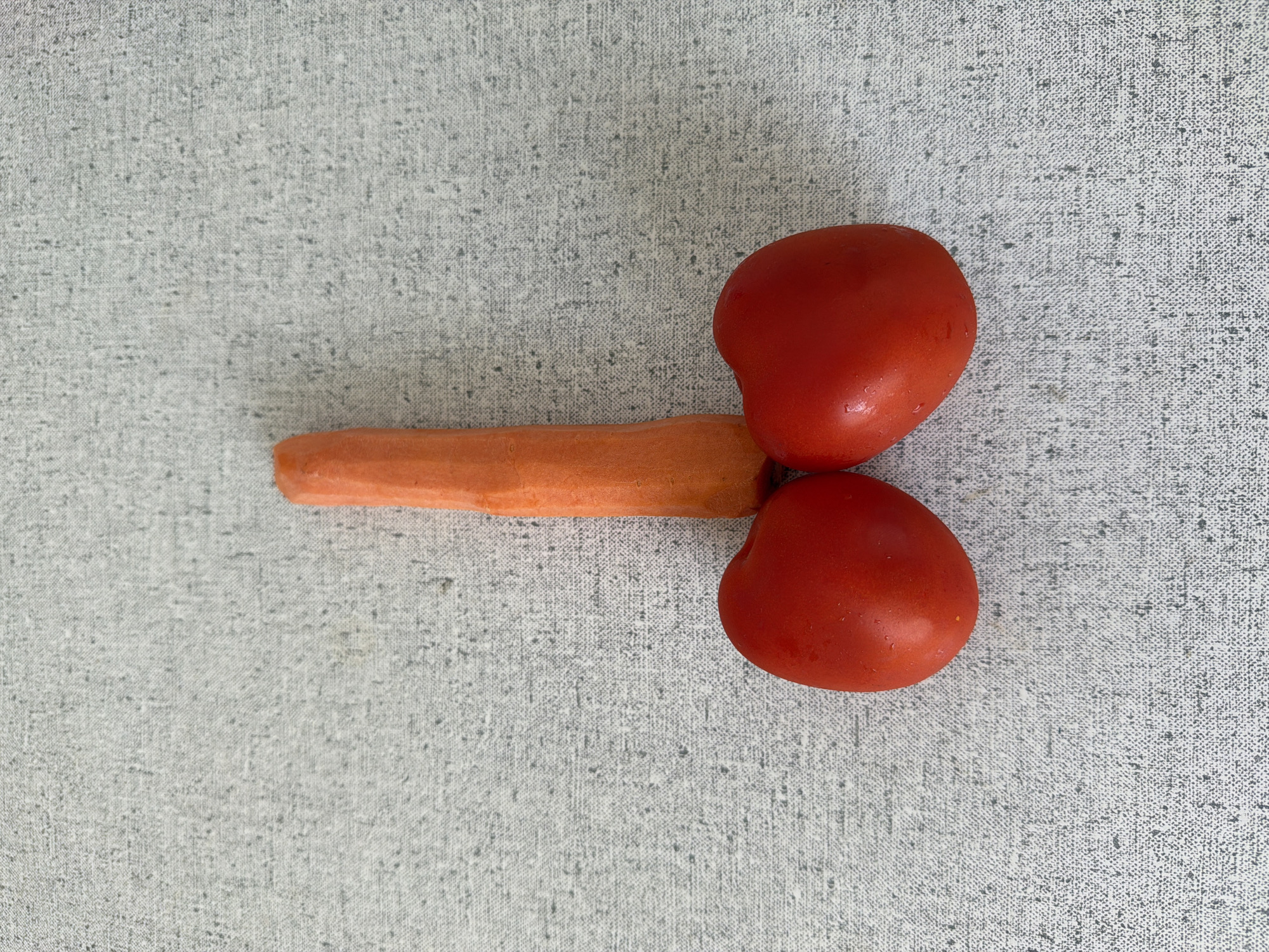 The image displays a carrot and two tomatoes arranged on a gray textured surface. The carrot is positioned horizontally between the two tomatoes, which are placed on top of each other. There are no visible people or indications of location, so identifying Moscow, Russia is not possible.