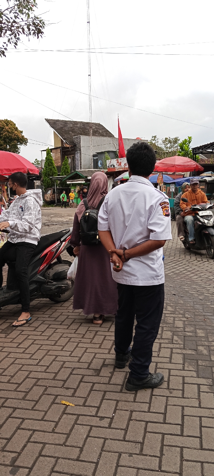 The image depicts a street scene in Banjaran, Indonesia (latitude: -7.046, longitude: 107.581). A man in a white shirt with a "POLDA" patch, black pants and black shoes is standing in the foreground. He has his hands clasped behind his back. In the background, a woman in a brown hijab and long brown skirt is standing. She is wearing a brown backpack and holding a white plastic bag. To the left of the image, another man, wearing a gray sweatshirt, black pants, and sandals, is standing next to a red and black motorcycle. He is holding a black helmet and a red and black object. In the background, there is a large metal tower, a multi-story building, and a street market with several stalls selling goods. There are also multiple people walking in the background. The street is paved with gray interlocking bricks, with some small green plants and a yellow leaf scattered on the ground. The sky is overcast and gray, with the sun obscured by clouds.