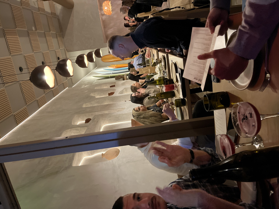 The image captures an indoor scene, likely a restaurant or bar, illuminated by warm, ambient lighting. The perspective is from a table, looking across a dining area where several groups of people are seated. The overall impression is that of an evening gathering or dinner.
In the immediate foreground, a man with dark hair and a beard is partially visible on the left, wearing a plaid shirt. His left hand is raised, possibly in a gesture or holding something out of frame. His face is turned towards the camera. On the right side of the foreground, parts of a table are visible with a dark wine bottle and two wine glasses containing red wine.
Further into the scene, a long table extends from left to right, where multiple individuals are seated. Closest to the foreground man, a person with long blonde hair is visible, facing away from the camera. Next to them, a woman with dark hair and glasses is also seated. Other diners are visible further down the table, engaged in conversation or looking around. A server, identified by a light blue long-sleeved shirt, is seen on the far right, holding an open menu with their left hand and pointing with their right index finger towards the table, suggesting they are assisting diners or taking an order. The words "del mar" are clearly visible at the bottom of the menu.
The restaurant's interior design features a textured wall on the left side with a pattern of horizontal slats. Hanging from the ceiling along this wall is a row of modern pendant lights, each featuring a dark, bowl-shaped fixture with an exposed, glowing bulb emitting a warm light. On the opposite wall, several decorative surfboards in varying colors (light wood, blue, green) are mounted horizontally, adding a distinct coastal or beach-inspired theme. Below the surfboards, the wall is adorned with sculpted, wave-like alcoves or niches, some containing small, light-colored pottery. Integrated strip lighting runs along the top edges of these architectural features, providing additional soft illumination. The tables are set with plates, cutlery, and several green wine bottles, some appearing empty, others possibly holding candles or flowers. In the background, more tables and diners are discernible, along with a distinct, woven light fixture hanging from the ceiling. The walls throughout the establishment appear to be a light, earthy tone, possibly concrete or stucco, contributing to a contemporary and inviting atmosphere.