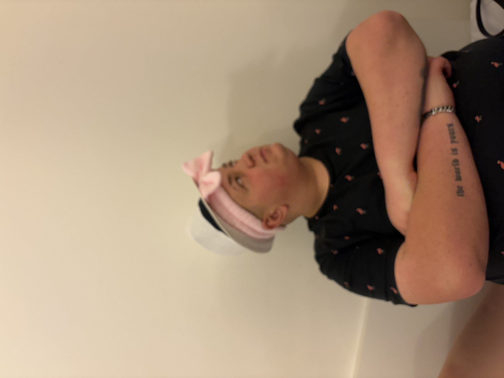 The image shows a person with light skin and short hair wearing a white sailor cap with a pink bow. They are also wearing a black shirt with small red floral patterns. The person has their arms crossed in front of them, and there is a tattoo on their right forearm that reads "the world is yours." The background is a plain, off-white wall. There is no clear indication of this image being in Jesolo, Italy or any other specific location.