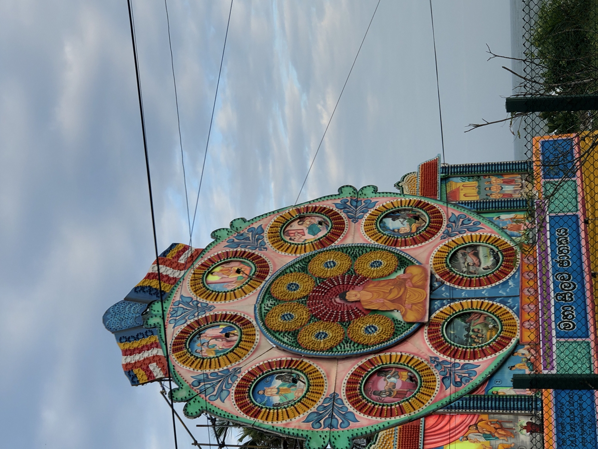 This image captures a close-up view of an ornately decorated structure, likely a festival float or temple decoration, against a cloudy sky. The central subject is a large, circular, multi-layered artistic piece adorned with numerous smaller circular panels, each containing painted scenes with figures. The dominant colors are pastel shades of pink, green, yellow, and blue, with intricate details and patterns. A prominent scene in the center depicts a figure in a saffron robe, possibly a religious icon. Other circular panels show various human figures in stylized settings.
The structure itself is elaborately designed with a scalloped edge and appears to be constructed from wood or a similar material, painted with vibrant imagery. To the right of the central piece, a vertical section of the decoration is visible, featuring rows of painted figures and what appears to be lettering in a non-Latin script. A green wire fence runs along the right edge of the frame, partially obscuring the structure and revealing a hint of greenery and a distant body of water or hazy landscape in the background. Electrical wires crisscross the sky, suggesting an outdoor setting. The lighting suggests it is daytime, with diffused light from the overcast sky. The overall impression is one of a colorful and culturally significant celebration or display. The visible text on the vertical panel is in Sinhala script.