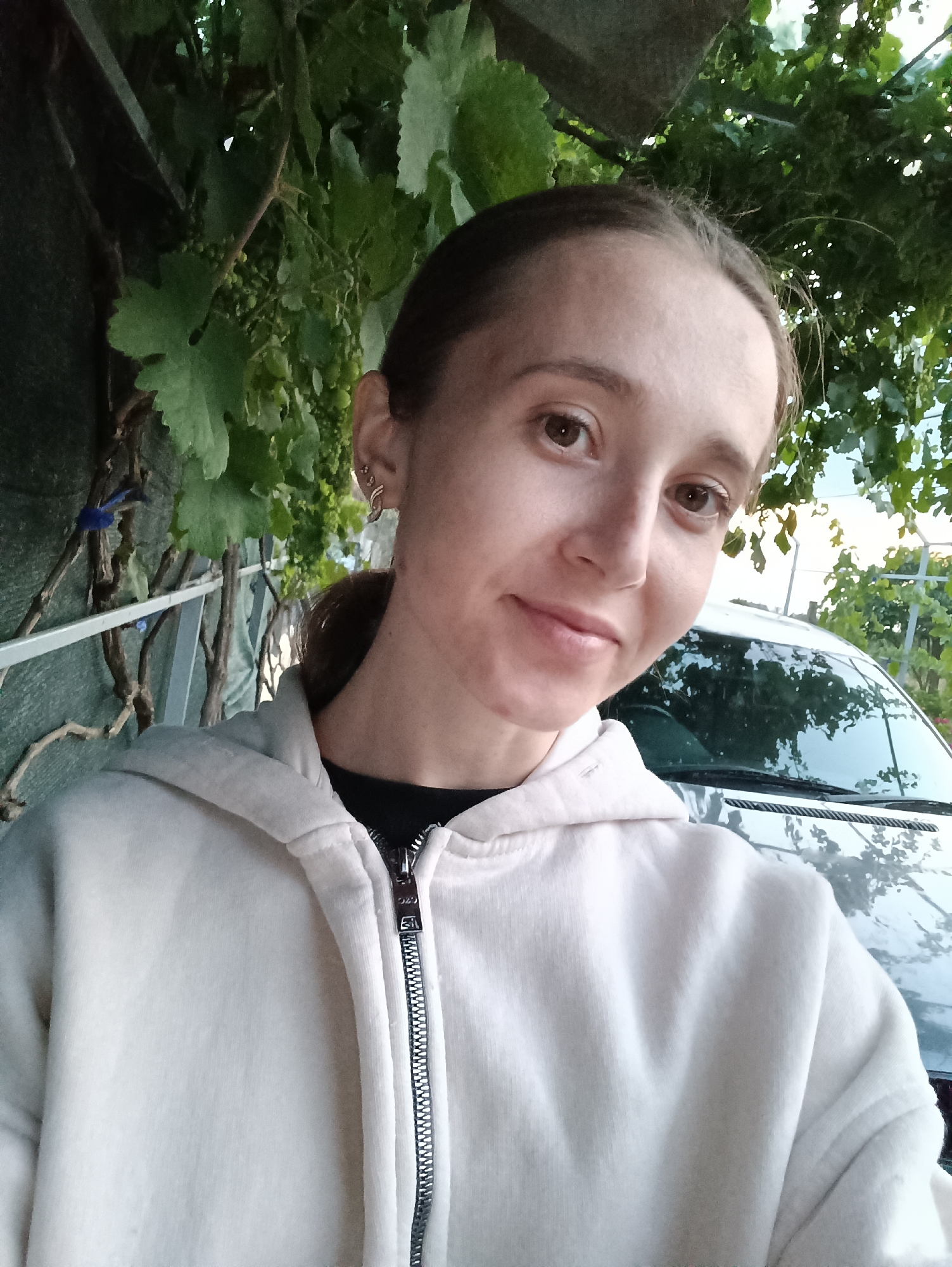 A young woman with light brown hair pulled back is taking a selfie outdoors. She is wearing a light-colored zip-up hoodie over a dark top. She has brown eyes and is smiling slightly. Behind her, a car is visible, and lush green grapevines are overhead, suggesting a location with vineyards. A fence and a green barrier are also present in the background, hinting at a residential or semi-rural setting. The image appears to be taken during the daytime. The location is identified as Vypasne, Ukraine.