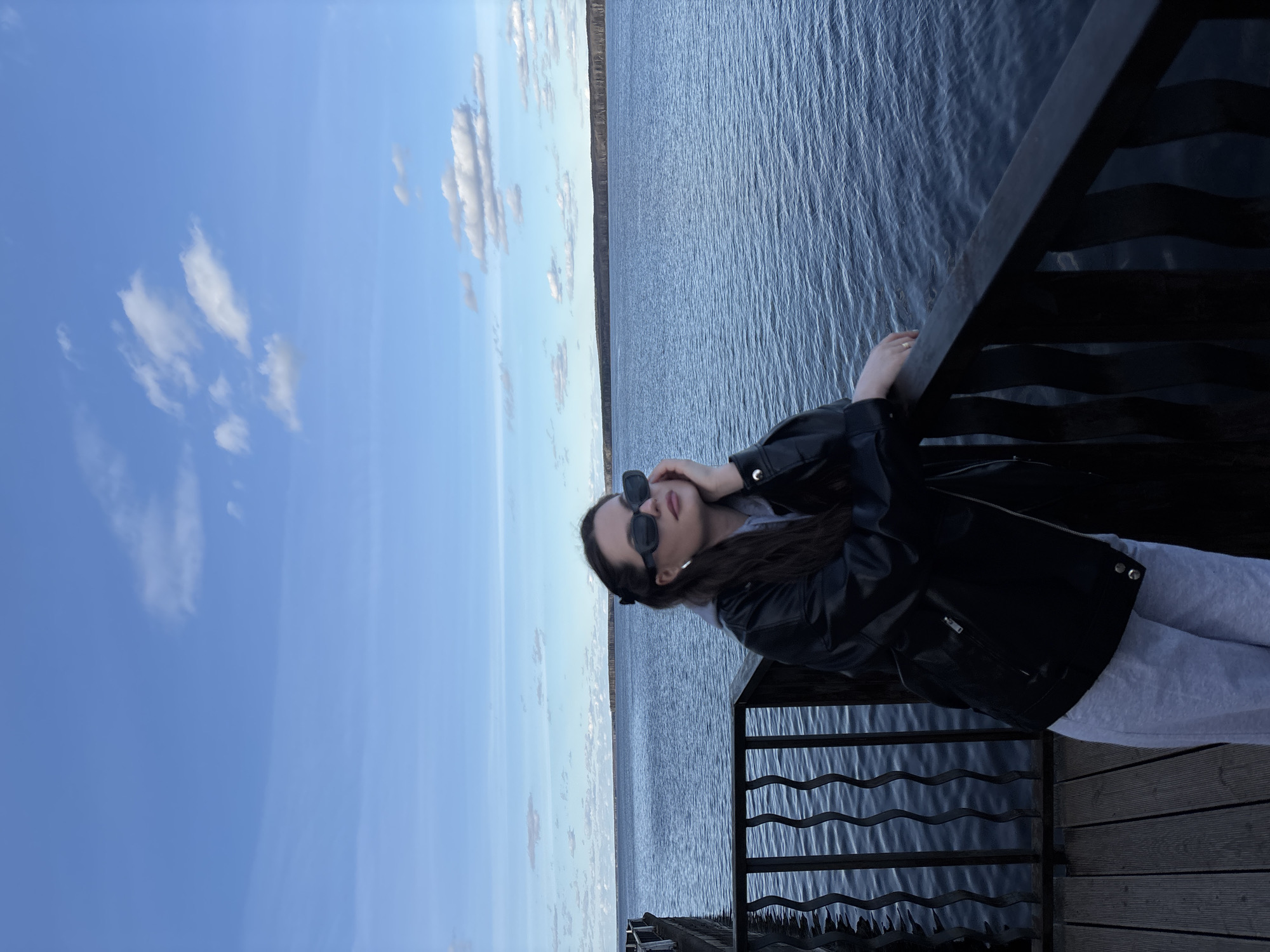 A woman stands on a wooden dock railing with a lake and skyline in the background. She wears sunglasses and a black leather jacket. The location is Chaikovsky, Russia, as evidenced by the landscape.