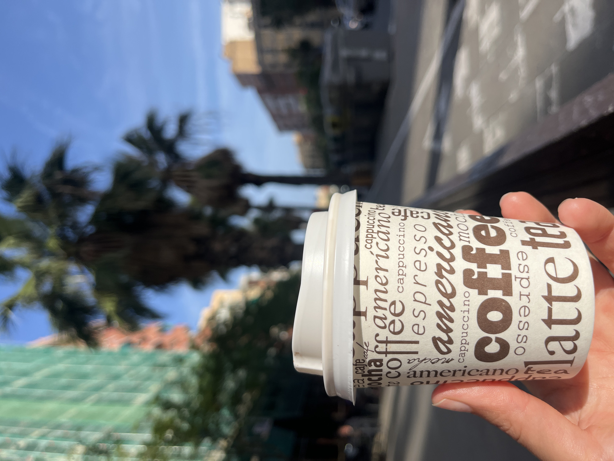 A first-person perspective shows a hand holding a white paper coffee cup with a white plastic lid. The cup is adorned with various coffee-related terms such as "coffee," "americano," "latte," "espresso," "cappuccino," "mocha," "tea," and "cafe," printed in brown text of different fonts and sizes.
The background is blurred, indicating a shallow depth of field. To the left, several large palm trees are visible against a clear, bright blue sky, suggesting sunny weather. Further back, indistinct urban buildings, likely apartment blocks, can be seen, along with a blurry green structure that might be part of a building facade or construction. A sidewalk or street is subtly visible on the right side of the frame. The scene captures a moment of enjoying a beverage outdoors in an urban environment, indicative of a daytime setting in a city like Barcelona, Spain.