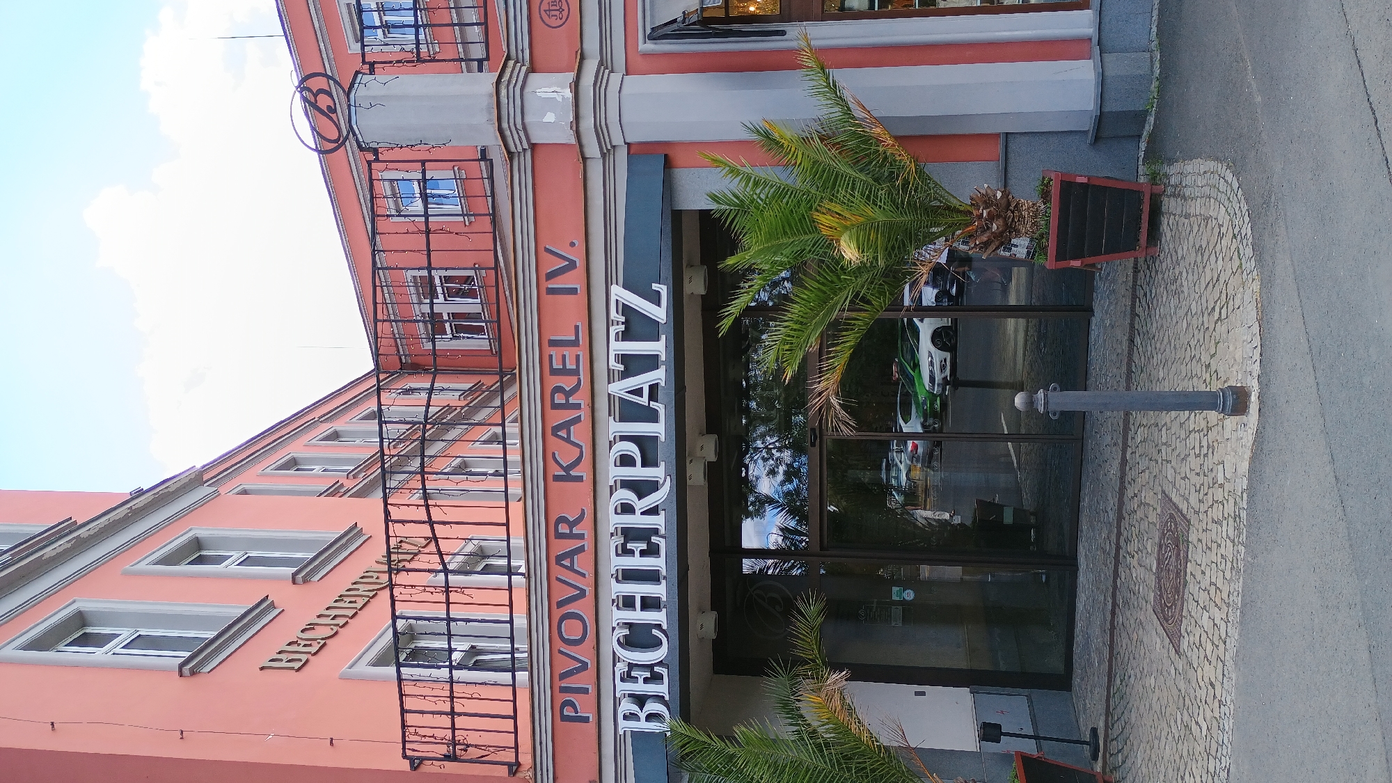 A wide shot captures the exterior of a building with a salmon-pink facade in Karlovy Vary, Czechia. The building features white-trimmed windows and a black wrought-iron balcony on the upper floors. A prominent sign above the entrance reads "PIVOVAR KAREL IV." in orange lettering, and below that, "BECHERPLATZ" in white capital letters on a dark background. The entrance is a large glass door. Palm trees in planters flank the entrance. A cobblestone pathway leads to the entrance, with a metallic bollard in the foreground. The sky above is a bright, hazy blue with scattered clouds. The overall impression is of a commercial establishment, likely a restaurant or brewery, in an urban setting.