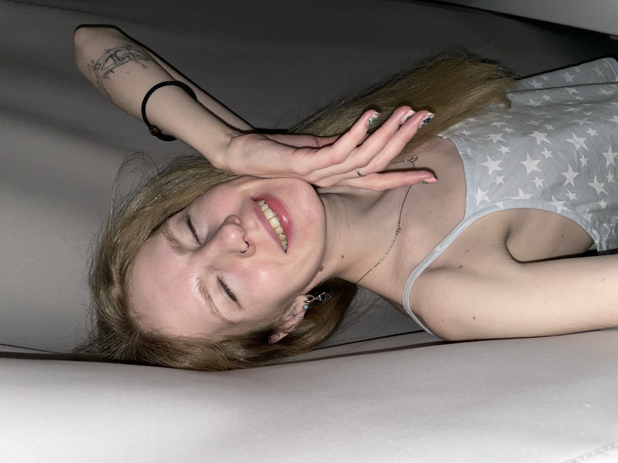 A young, fair-skinned woman with light blonde hair is lying on her side on a white surface. She is wearing a light blue tank top with white stars. Her right arm is raised above her head, with a black hair tie around her wrist and a tattoo on her inner forearm. Her left hand is near her face. She is smiling with her eyes closed and has a nose ring in her left nostril. The environment appears to be indoors, and the lighting is soft. The location is unknown and cannot be ascertained from the image provided.