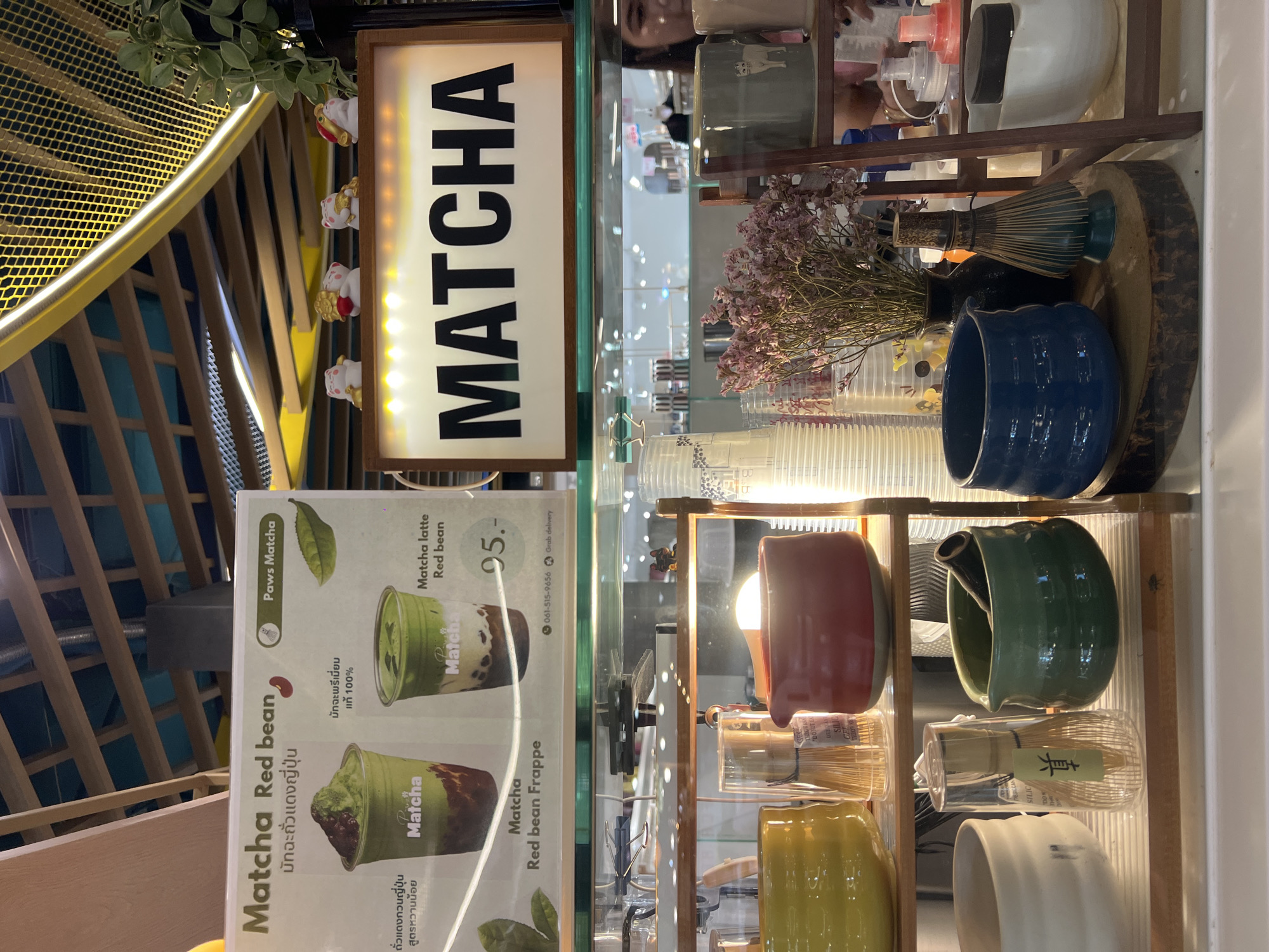 The image shows a display case with a menu for "Matcha Red Bean" drinks. The menu, titled "Paws Matcha," features descriptions and prices for Matcha Red Bean, Matcha Latte Red Bean (95.-), and Matcha Red Bean Frappe. The drinks are advertised as using premium matcha and red bean paste that is less sweet.
Behind the menu, the display case contains various matcha-related items. These include ceramic bowls in shades of blue, red, green, yellow, and white, as well as whisks for preparing matcha. There are also stacks of plastic cups and some dried flowers. A sign with the word "MATCHA" in bold, vertical lettering is visible above the menu.
The scene is set indoors, with a textured, yellow-toned structure and small lucky cat figurines above the display case. The image also captures a partial reflection of a person.