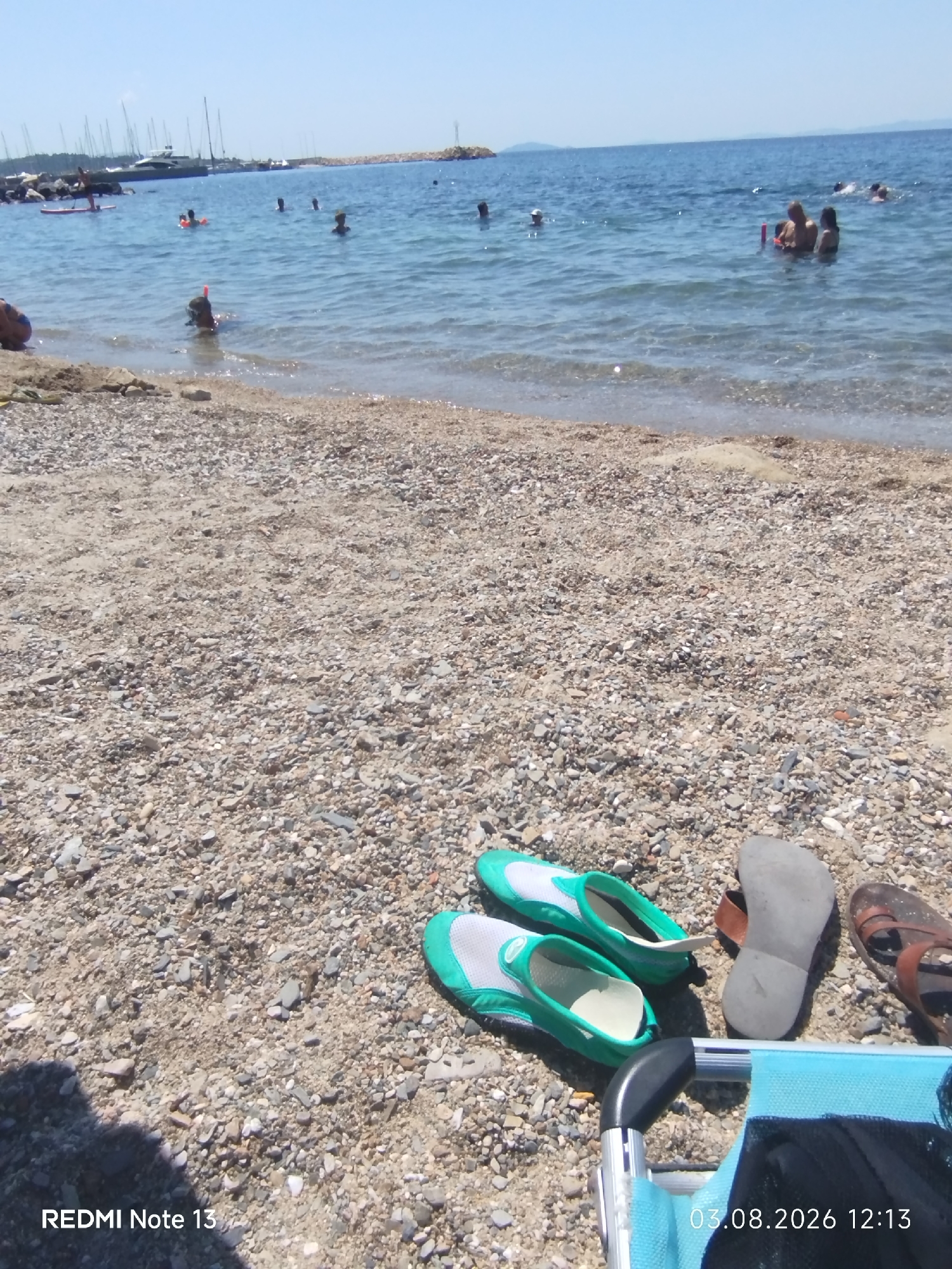 The image depicts a pebble beach in Sithonia Municipal Unit, Greece, on a clear, sunny day. The foreground is dominated by the textured beach, with a pair of green and white water shoes and two pairs of sandals placed on the pebbles. A portion of a blue beach chair is visible in the lower right corner. In the middle ground and background, the Aegean Sea is calm and blue, with several people visible swimming and wading. Some individuals are wearing snorkeling gear, and a boat with masts is anchored in the distance. The shoreline is lined with rocks and a breakwater. The image is dated August 3, 2026, at 12:13.